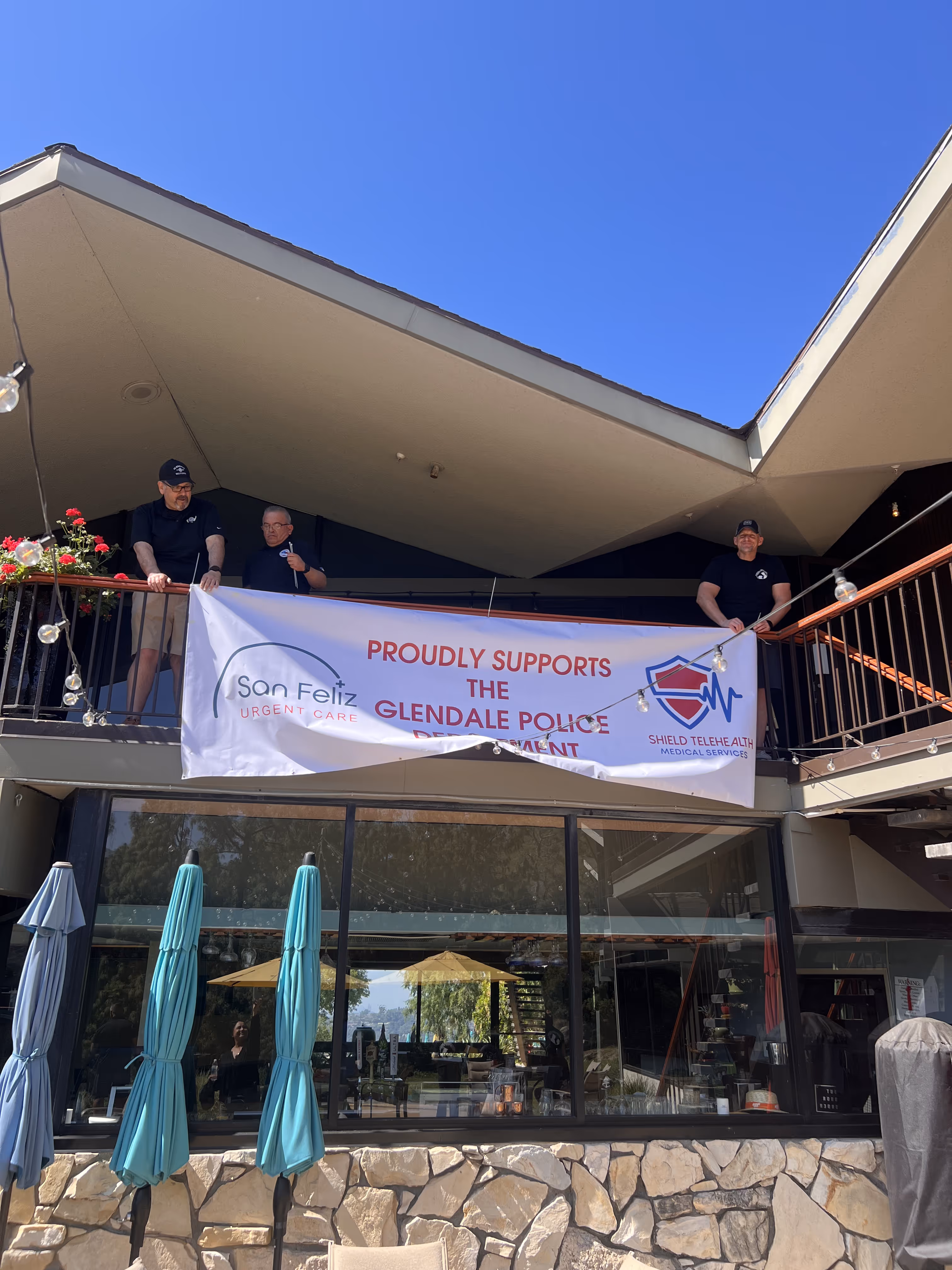 Three men on a balcony holding a banner that reads 'Proudly supports the Glendale Police Department' with logos for San Feliz Urgent Care and Shield Telehealth Medical Services.