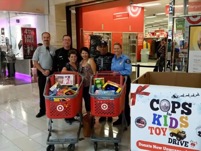 Group of people, including police officers and civilians, standing behind shopping carts filled with toys in a mall for a 'Cops for Kids Toy Drive' donation event.