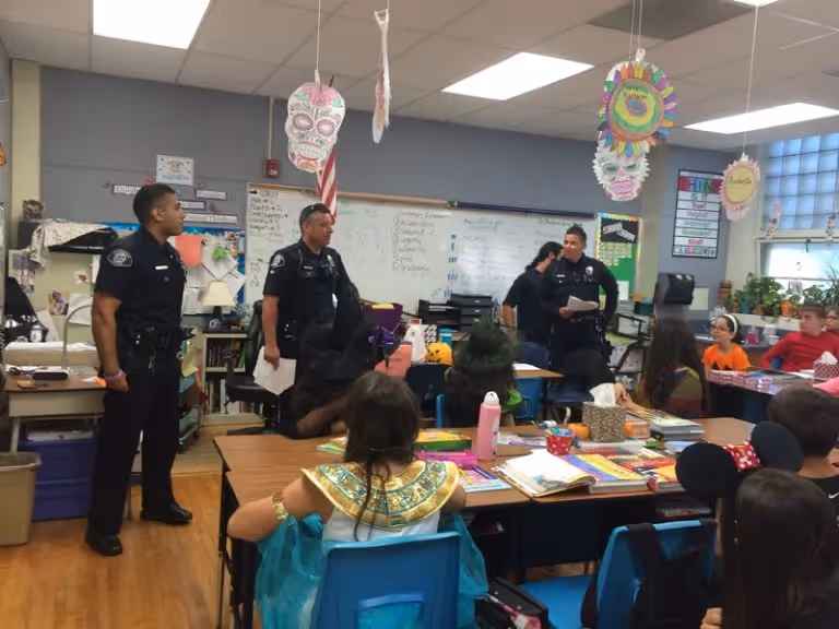 Three police officers standing and speaking to children in a classroom decorated for Halloween with colorful skulls and other festive decorations.