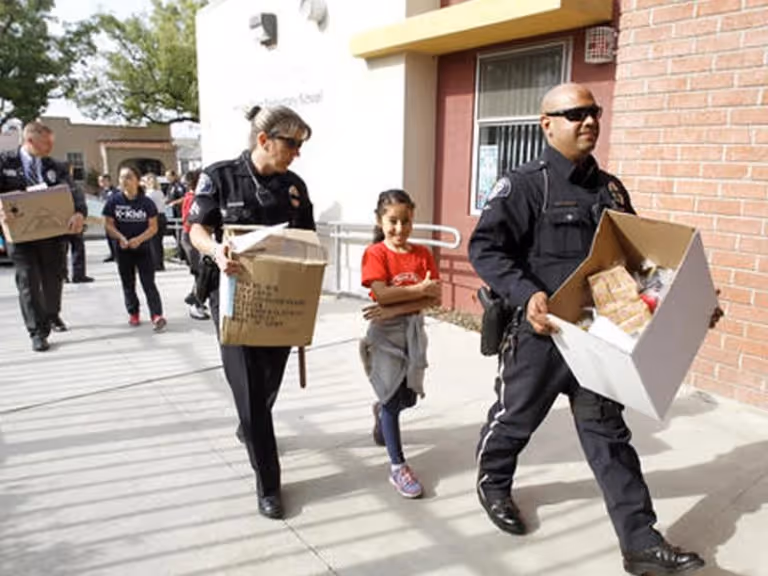 Two police officers and a young girl walking outside a building, each carrying boxes.
