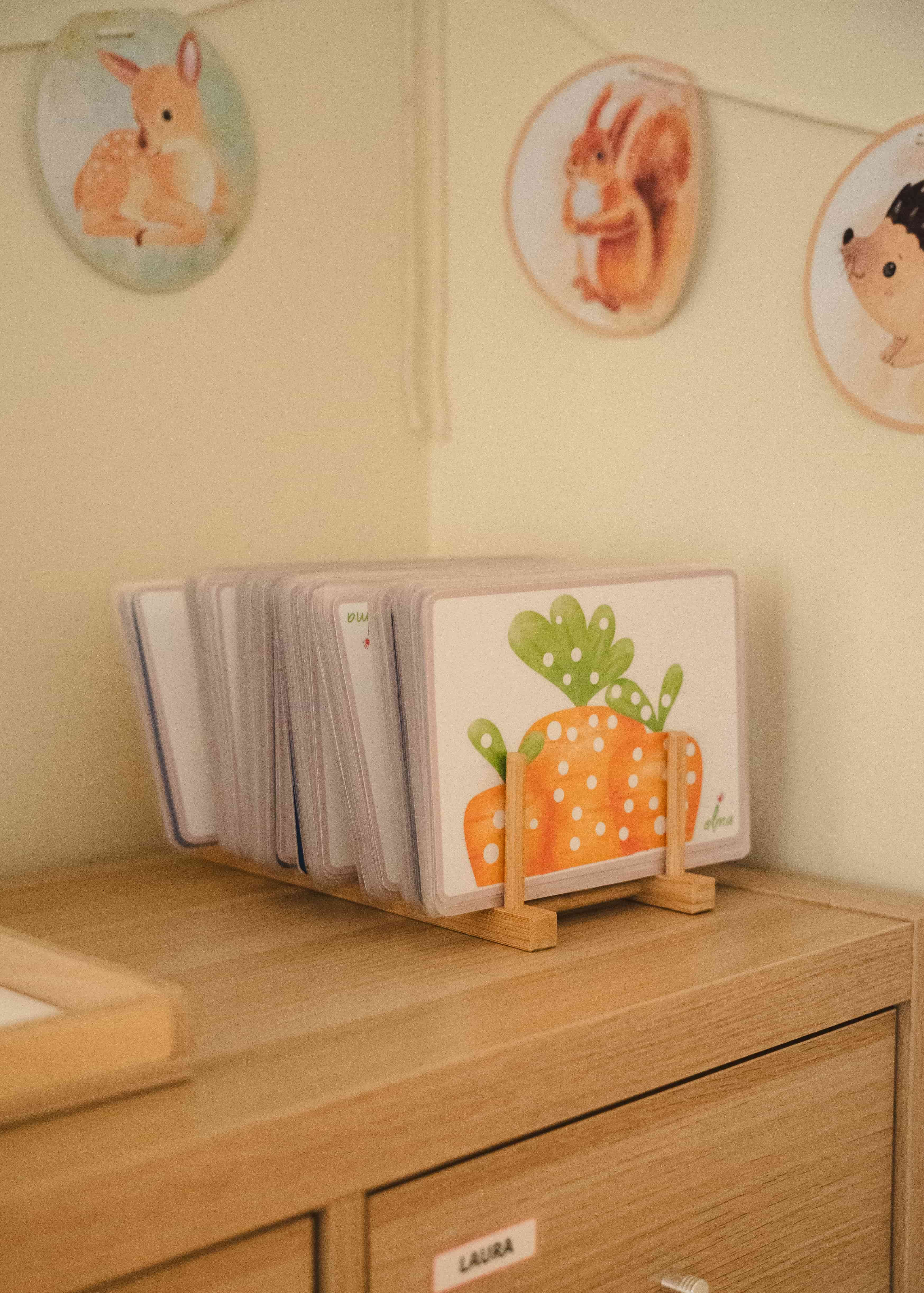 Koka plauktā uzglabātas bērnu kāršu spēles ar oranžas un zaļas burkāna ilustrāciju priekšpusē, fonā pie sienas redzamas dzīvnieku ilustrācijas.