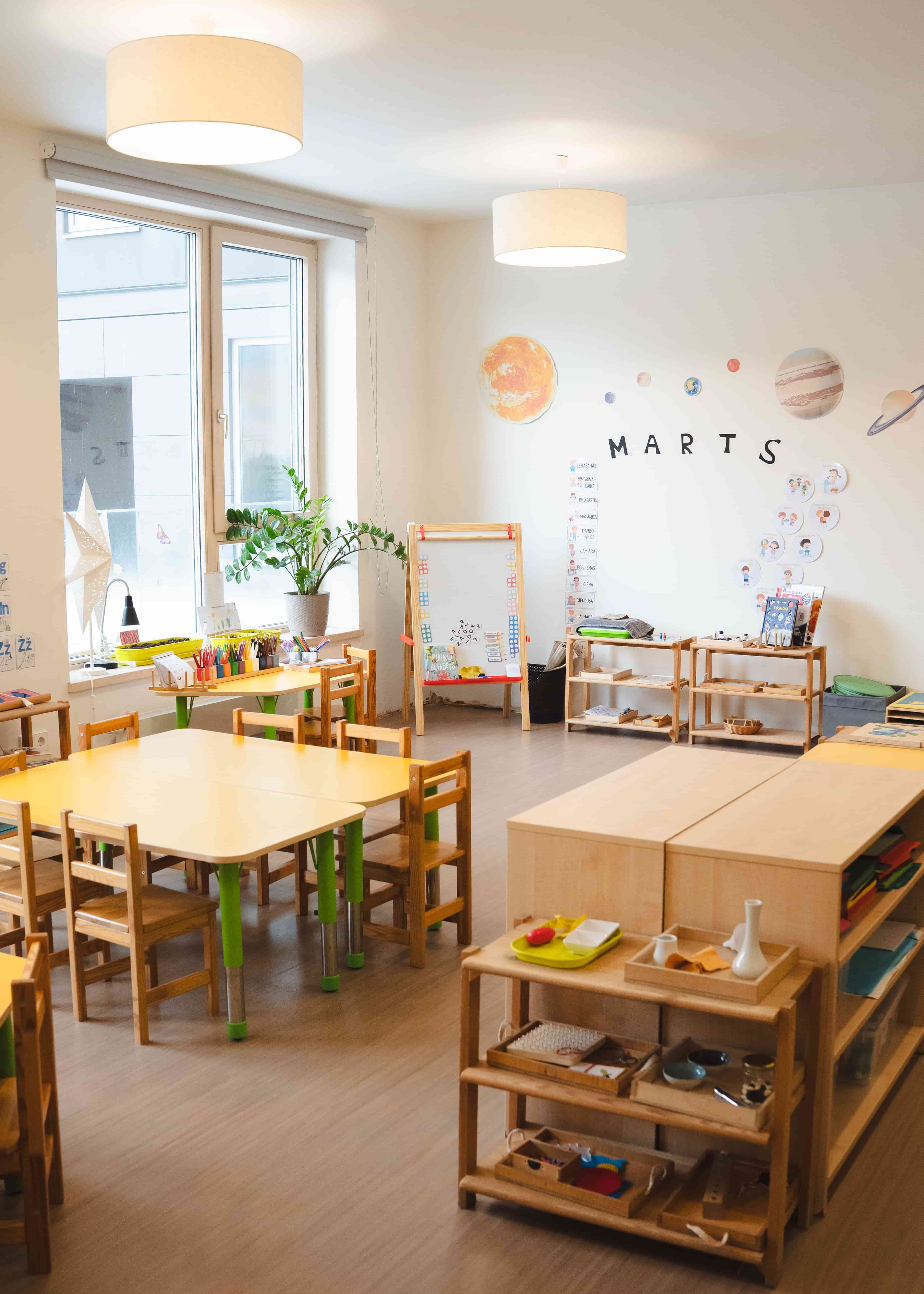 Krāsaina bērnu mācību telpa ar koka galdiem, krēsliem, zīmēšanas easel un sienas dekorācijām ar planētām un uzrakstu MARTS.