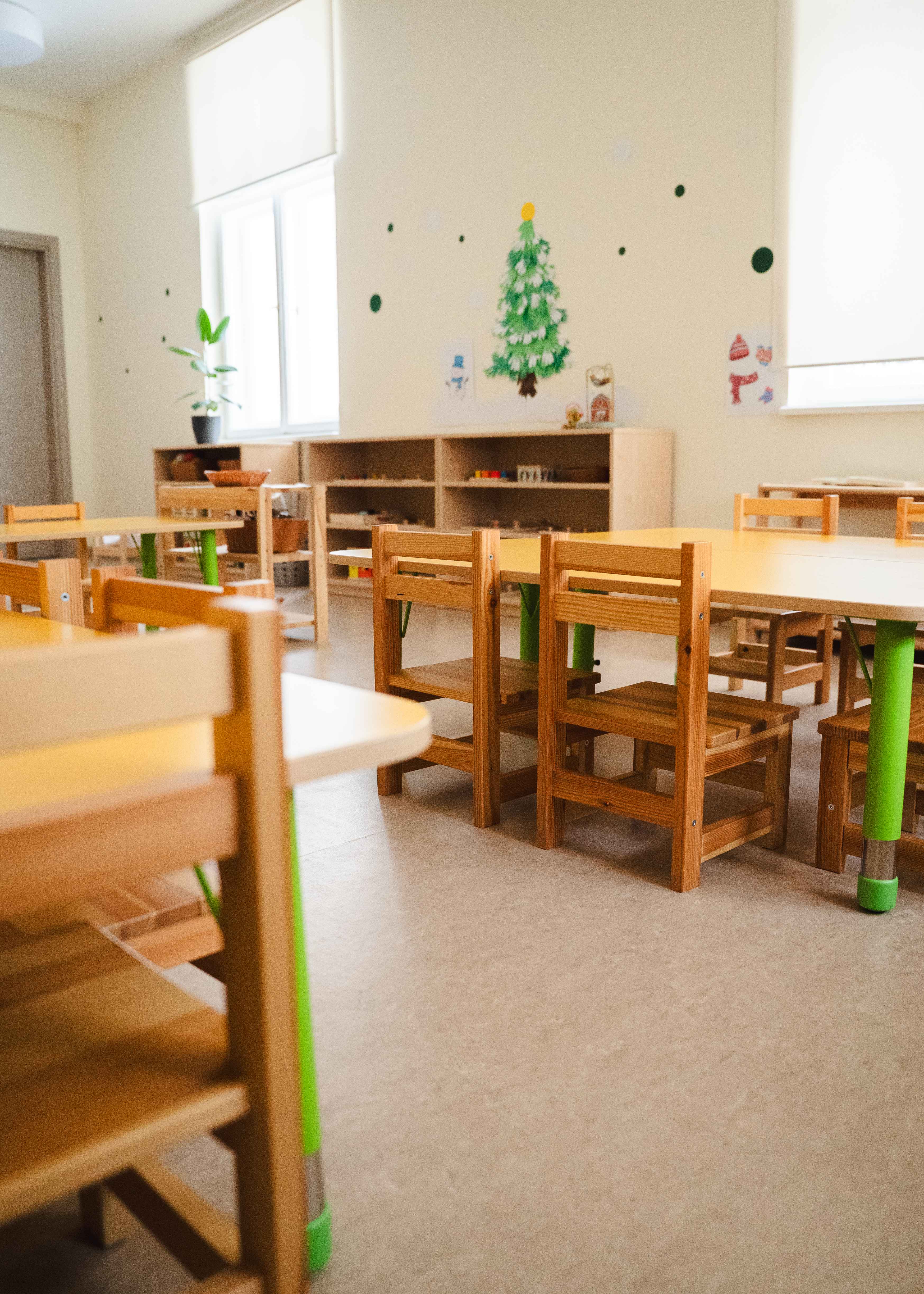 Klusa bērnudārza telpa ar koka krēsliem, galdiem un sienas dekorācijām, ieskaitot Ziemassvētku egli.