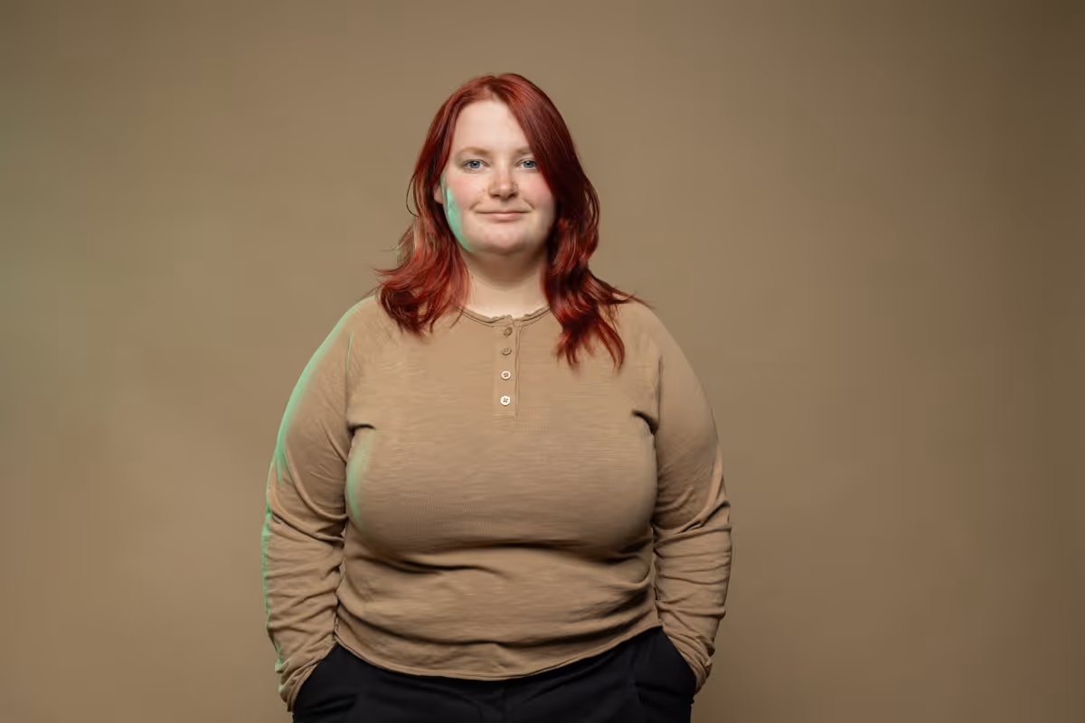 Frau mit roten Haaren, die ein beiges Langarmshirt und schwarze Hosen trägt, vor beigem Hintergrund.