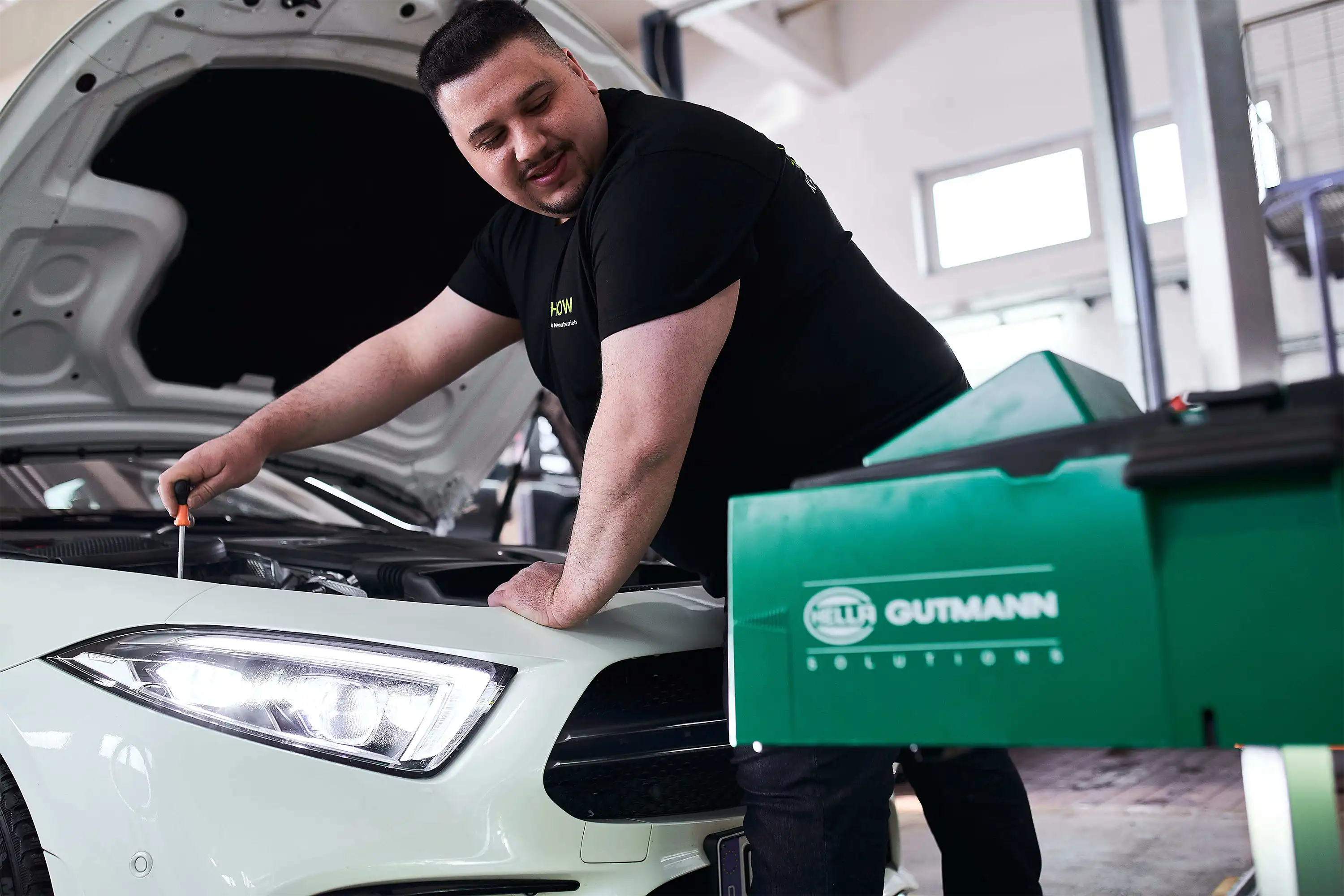 Mechanic with screwdriver working on a white car engine in a garage with green Hella Gutmann equipment.