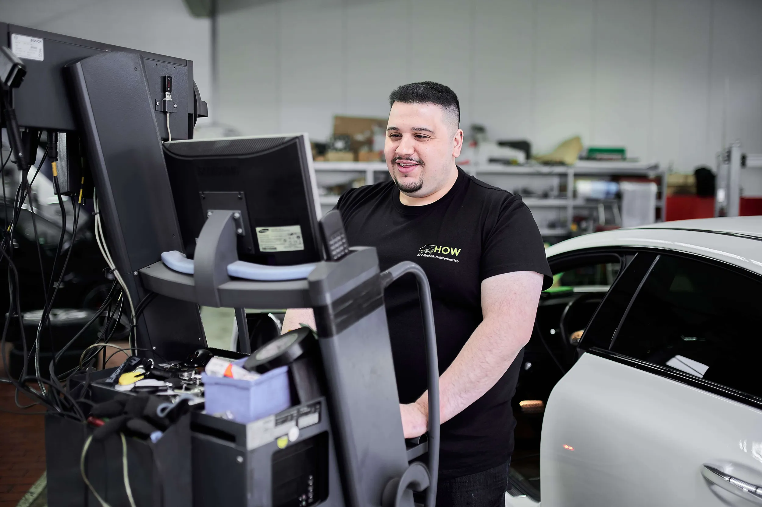 Man in a black HOW T-shirt smiling while using a diagnostic machine near a white car in a workshop.