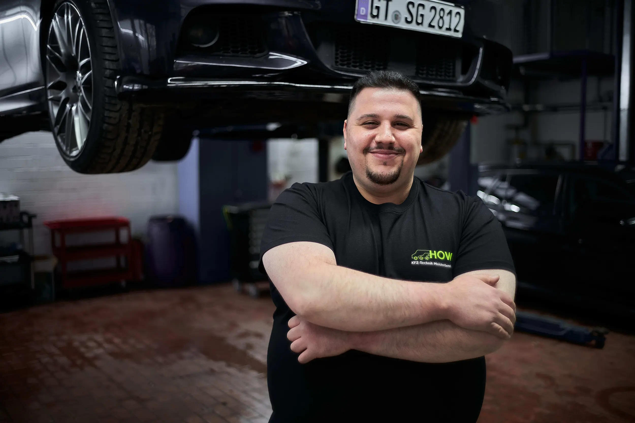 Smiling man with crossed arms in black shirt standing in auto repair shop under a car lift.