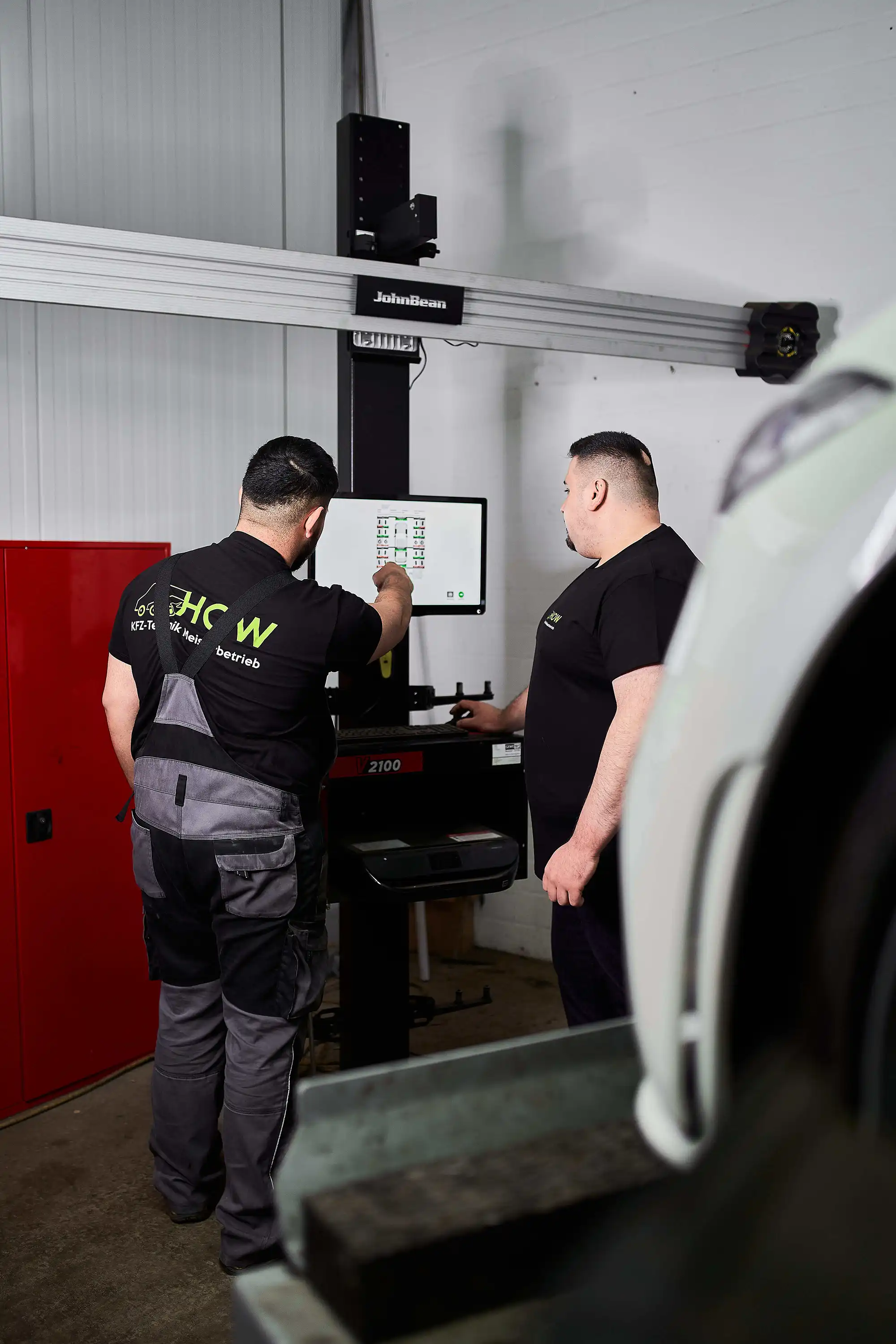 Two mechanics in black HOW shirts reviewing data on a John Bean automotive diagnostic machine.