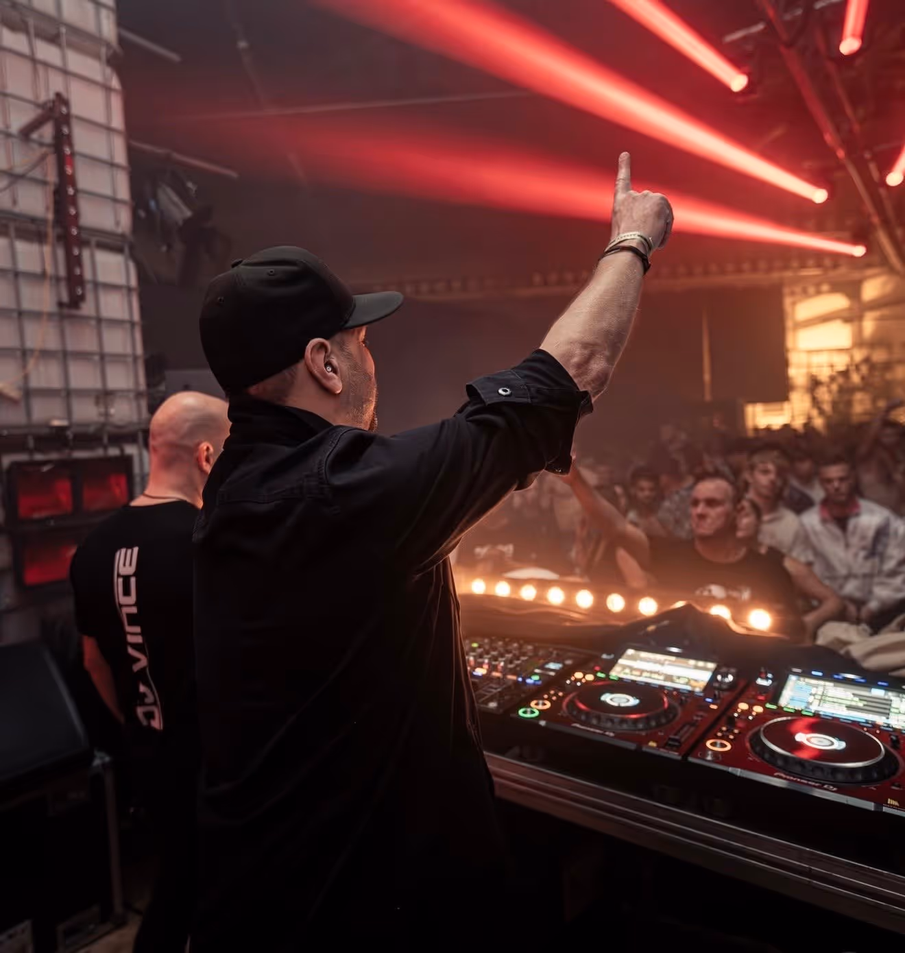 Dj die een vinger omhoog steekt tijdens een optreden in een club met rode verlichting en publiek.