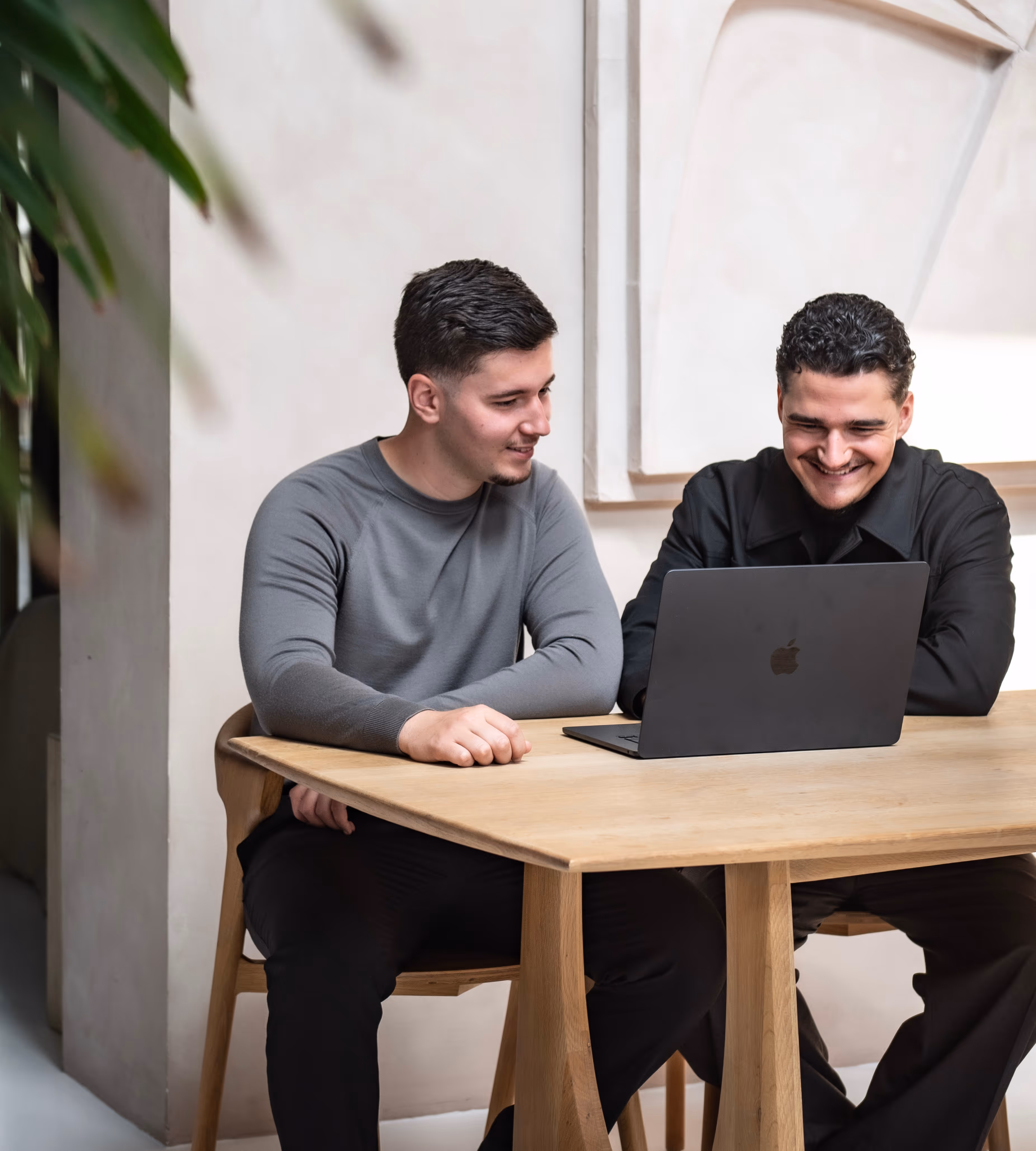 Twee jonge mannen zitten aan een houten tafel en kijken samen naar een laptop met Apple-logo.