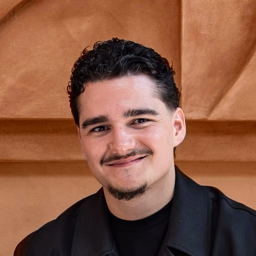Jonge man met donkere krullen en snor lacht vriendelijk naar de camera, met beige achtergrond.