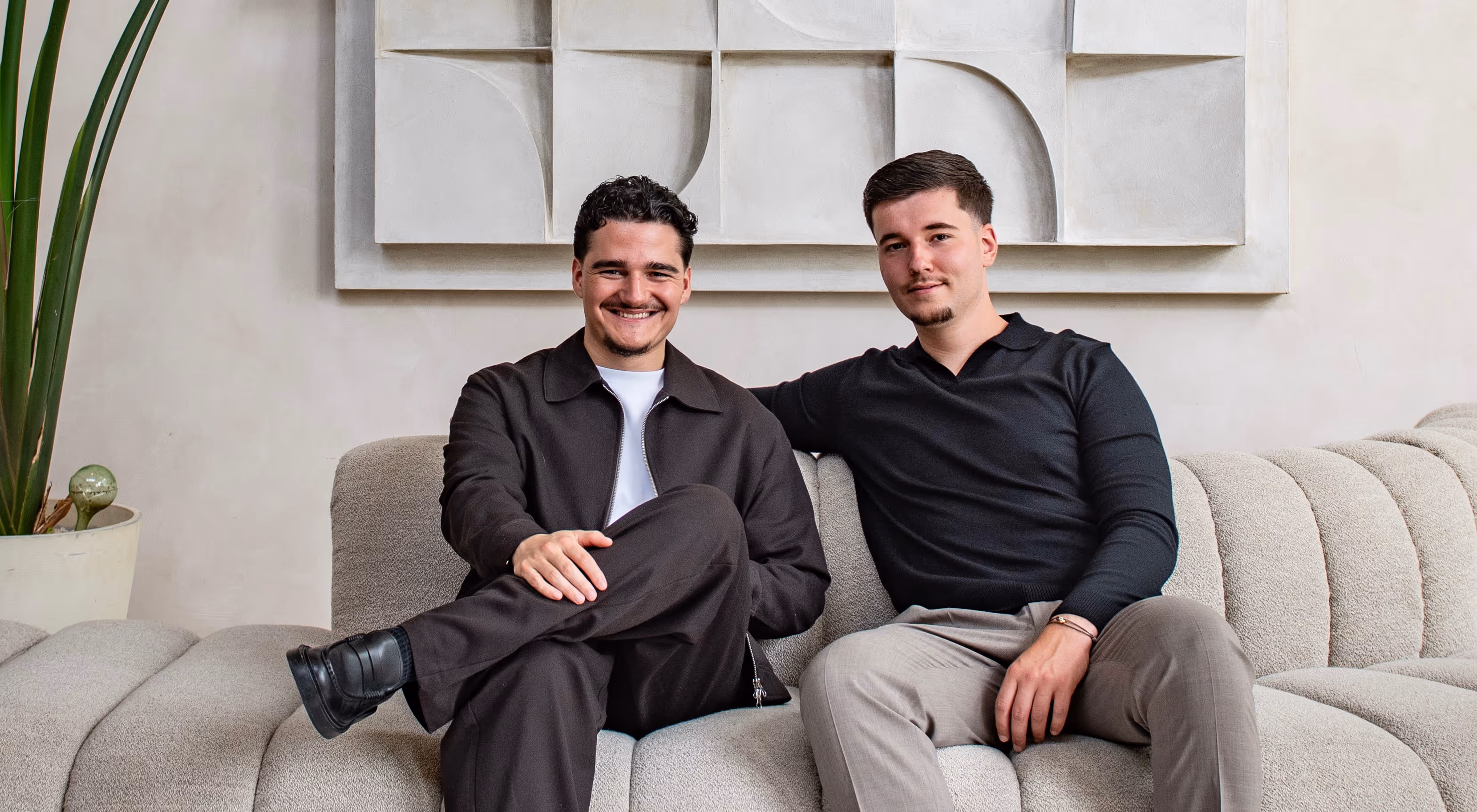 Twee jonge mannen zitten op een beige bank voor een muur met abstract kunstwerk.