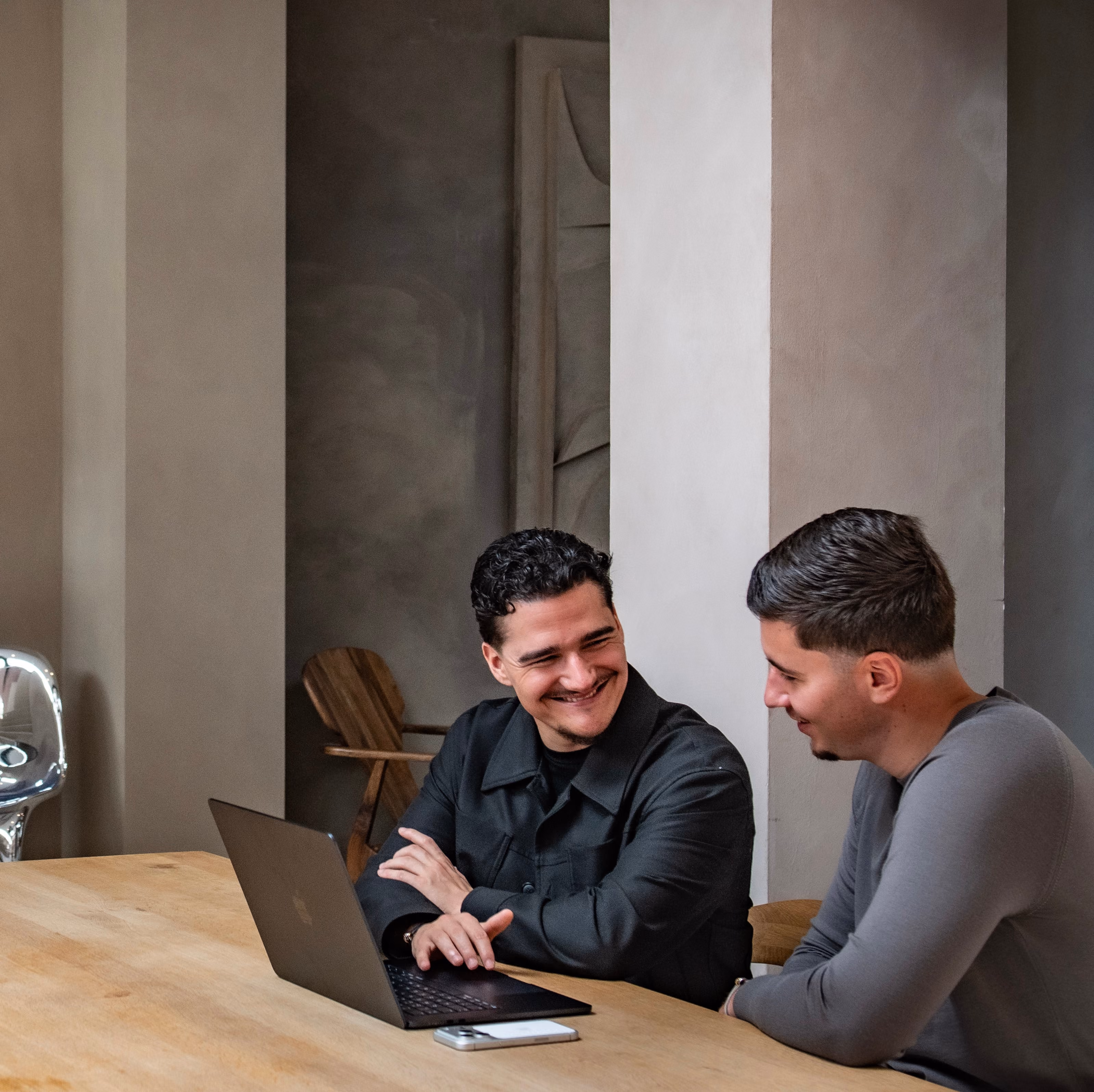 Twee mannen zitten aan tafel met een laptop en praten in een moderne neutrale ruimte.