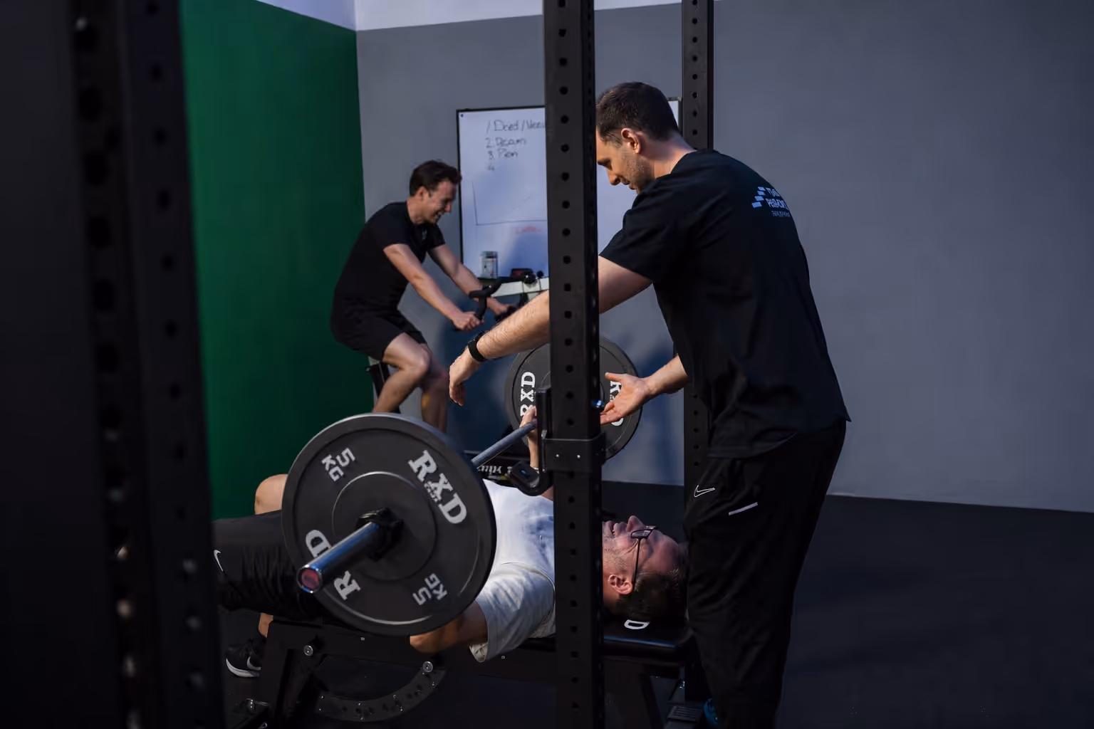 Man die een barbell-bankdrukapparaat gebruikt met begeleiding terwijl een ander man fietst in de sportschool.