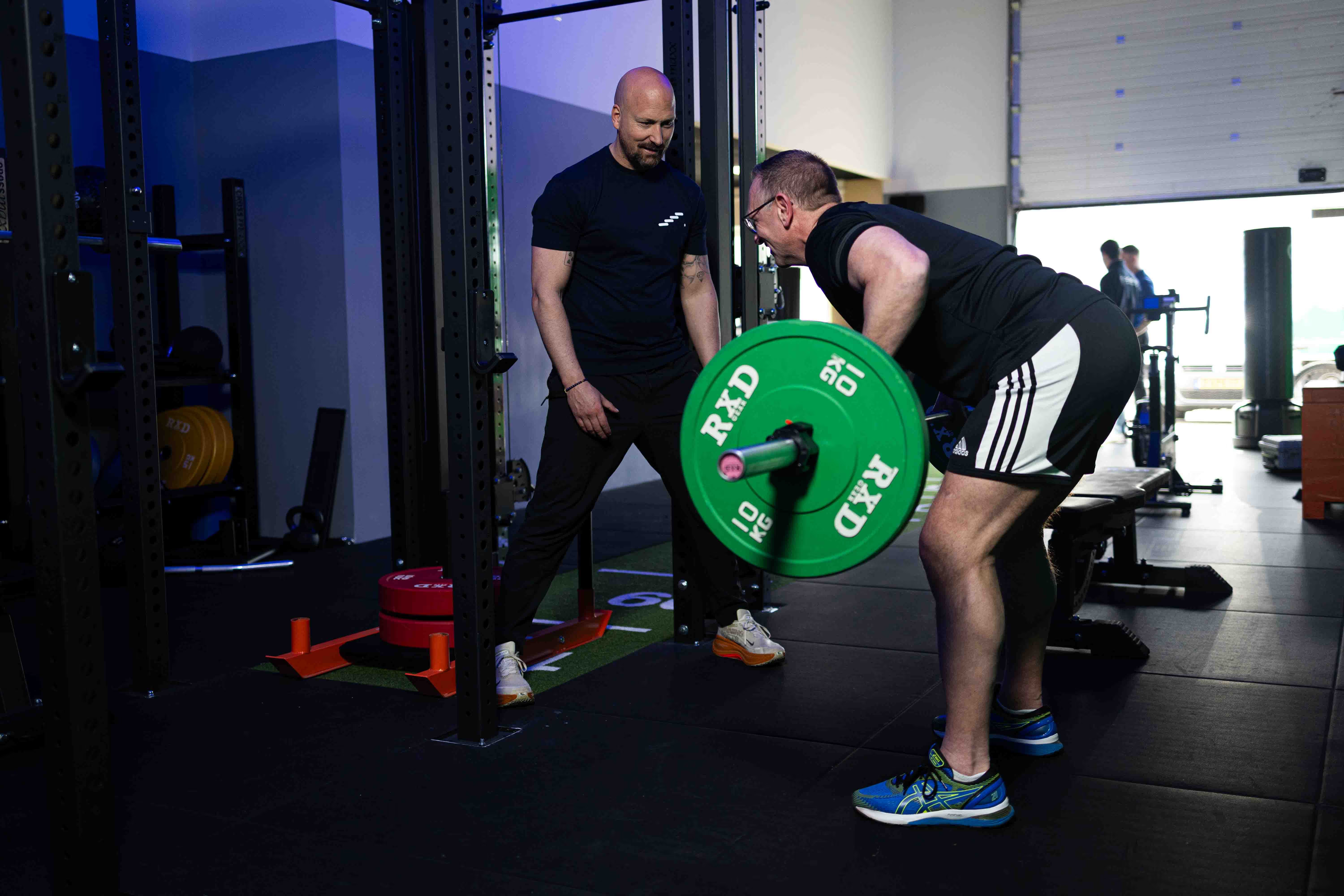 Man tilt naar voren tilt met gewichten terwijl een trainer toekijkt in een sportschool.