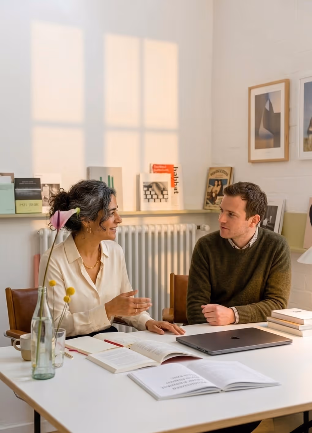Two people sit at a table with open books and laptop, engaged in a friendly conversation indoors.