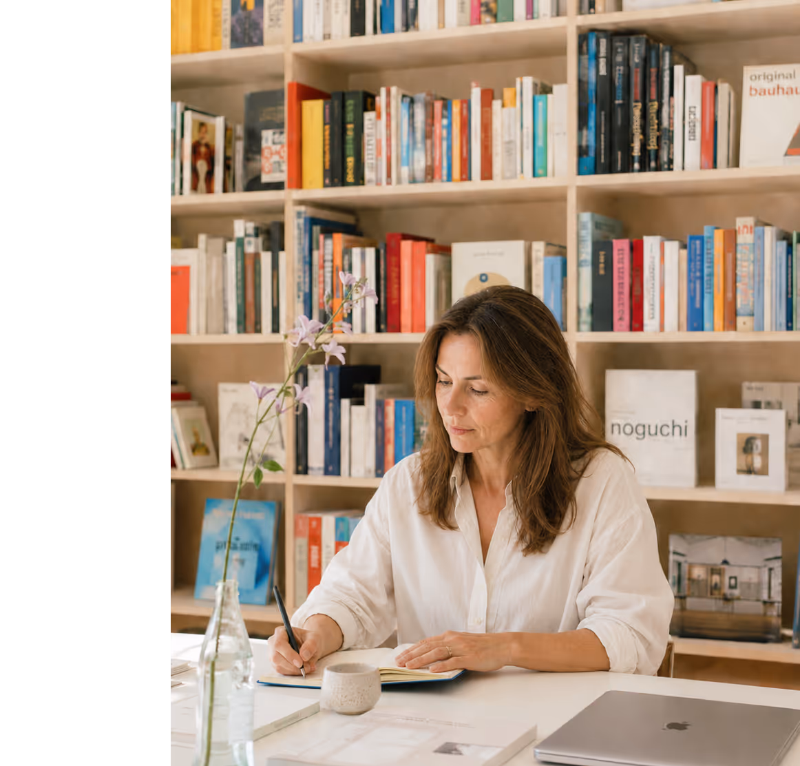Vrouw in wit overhemd schrijft in een notitieboek aan een bureau met boekenplank achter haar en laptop op bureau