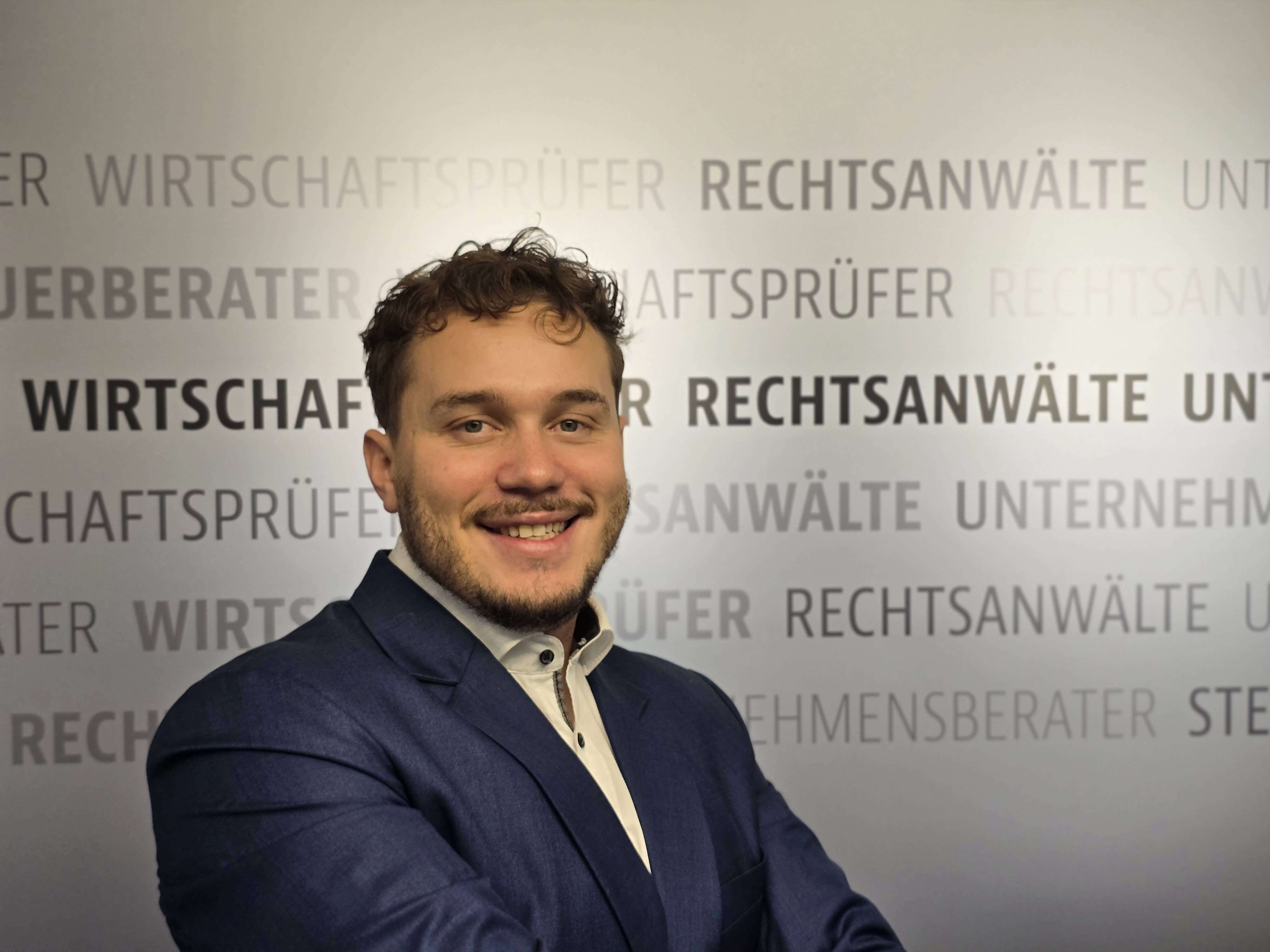 Rechtsanwalt im Anzug vor einer Kanzleiwand mit den Schriftzügen Rechtsanwälte, Wirtschaftsprüfer und Steuerberater