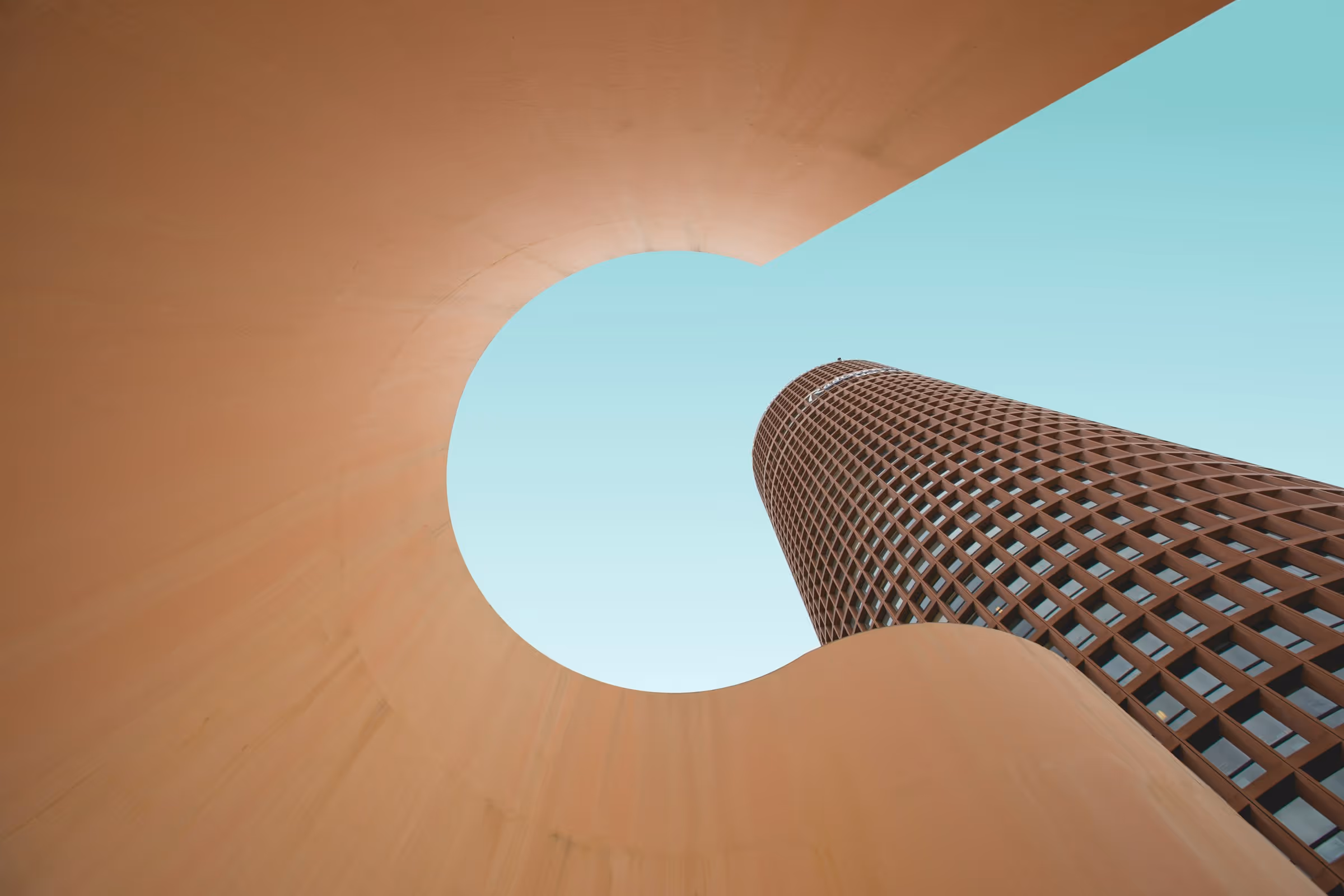 View looking up at a tall cylindrical building with a lattice facade, framed by a curved beige architectural structure against a clear blue sky.