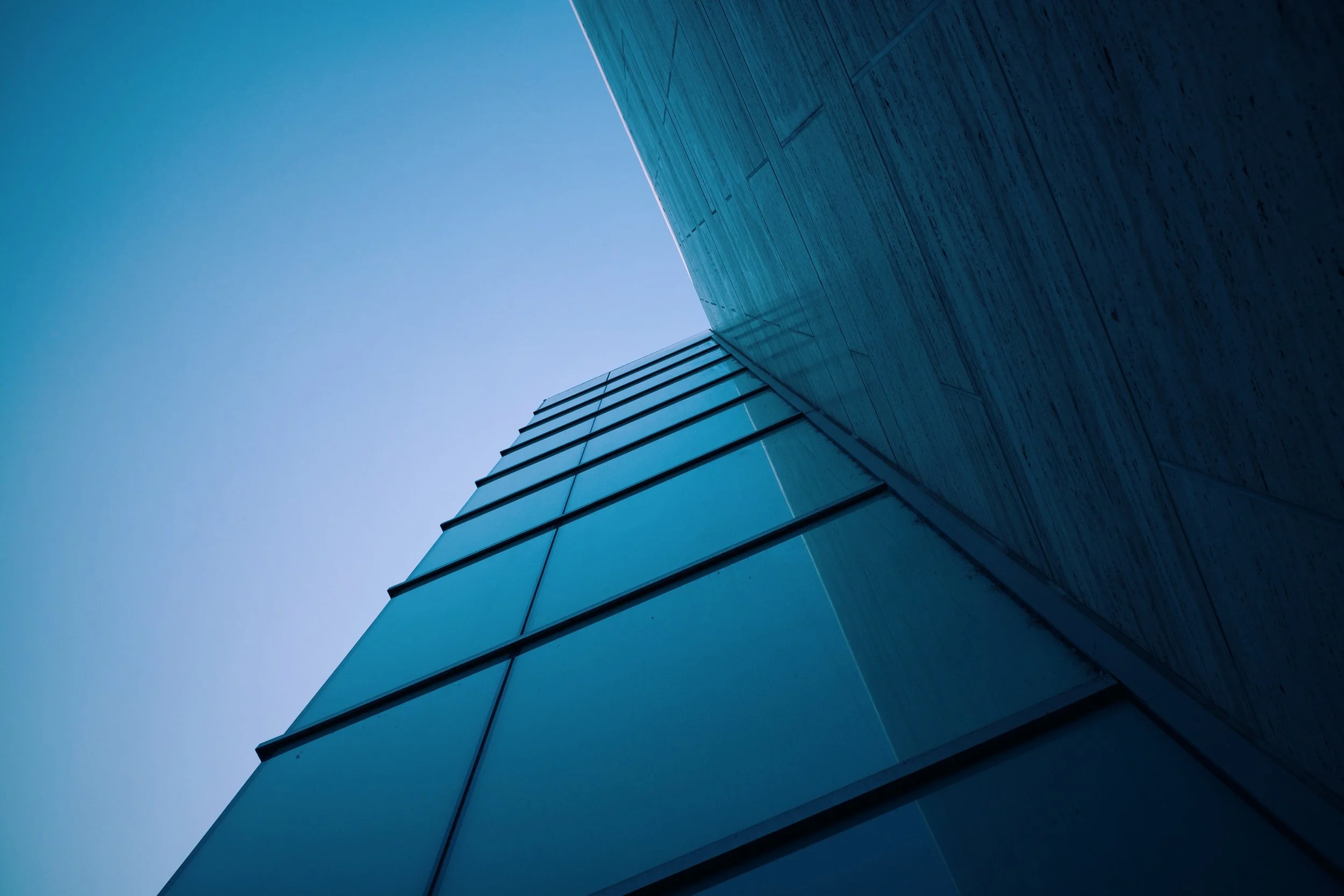 Low-angle view of a modern building with glass windows and a concrete wall against a clear blue sky.