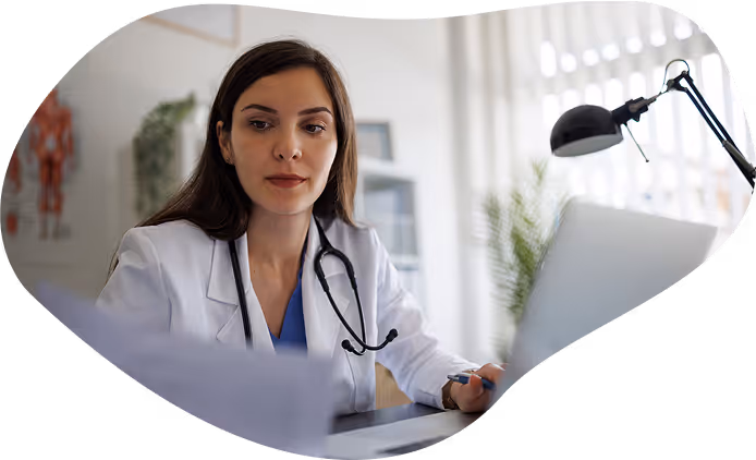 Female doctor with stethoscope reviewing documents in a bright office at a desk with a laptop.