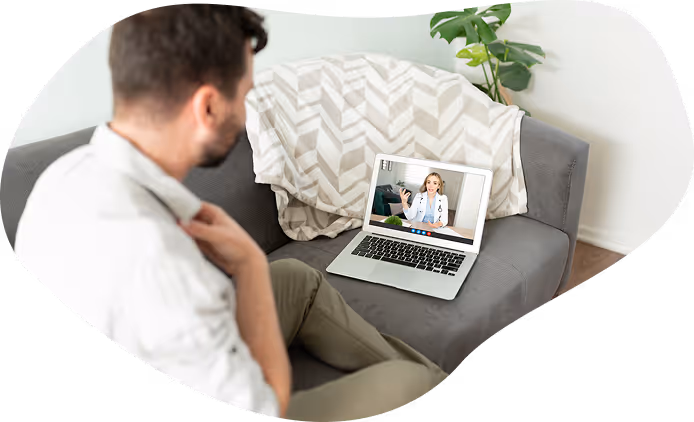 Man sitting on a couch having a telehealth video call with a female doctor on a laptop.