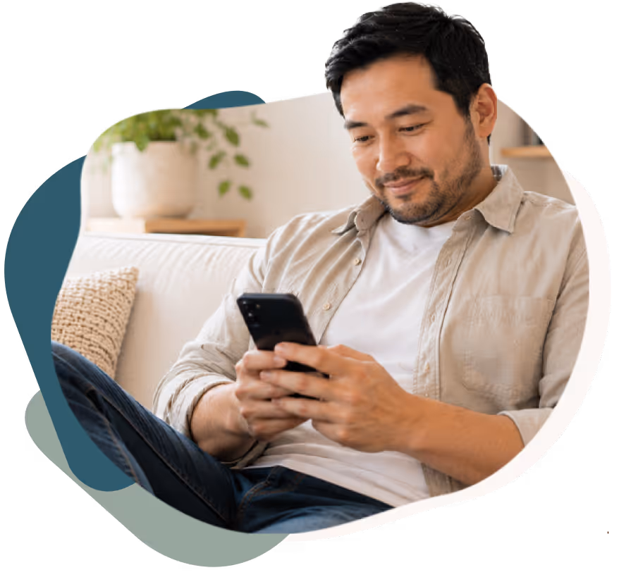 Smiling man sitting on a couch looking at his smartphone in a cozy living room.