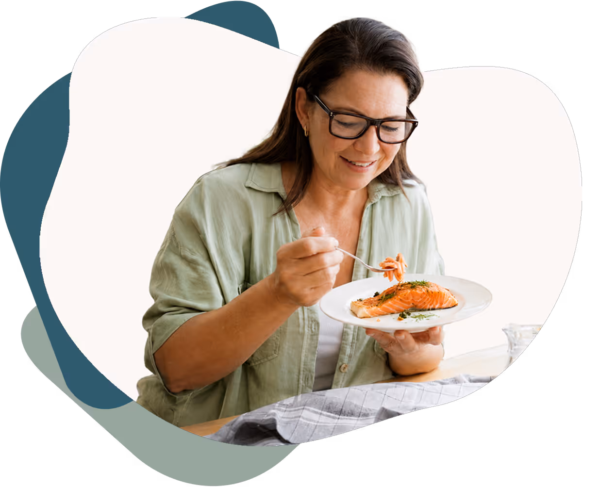 Smiling woman in glasses and green shirt eating a piece of cooked salmon on a white plate.