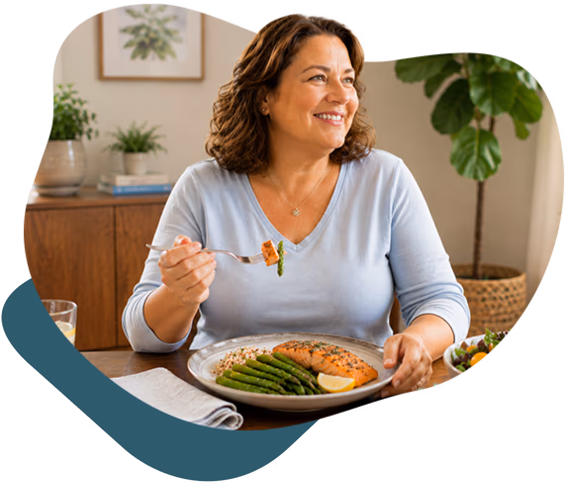 Smiling woman holding fork with salmon and asparagus, sitting at table with a meal and salad.