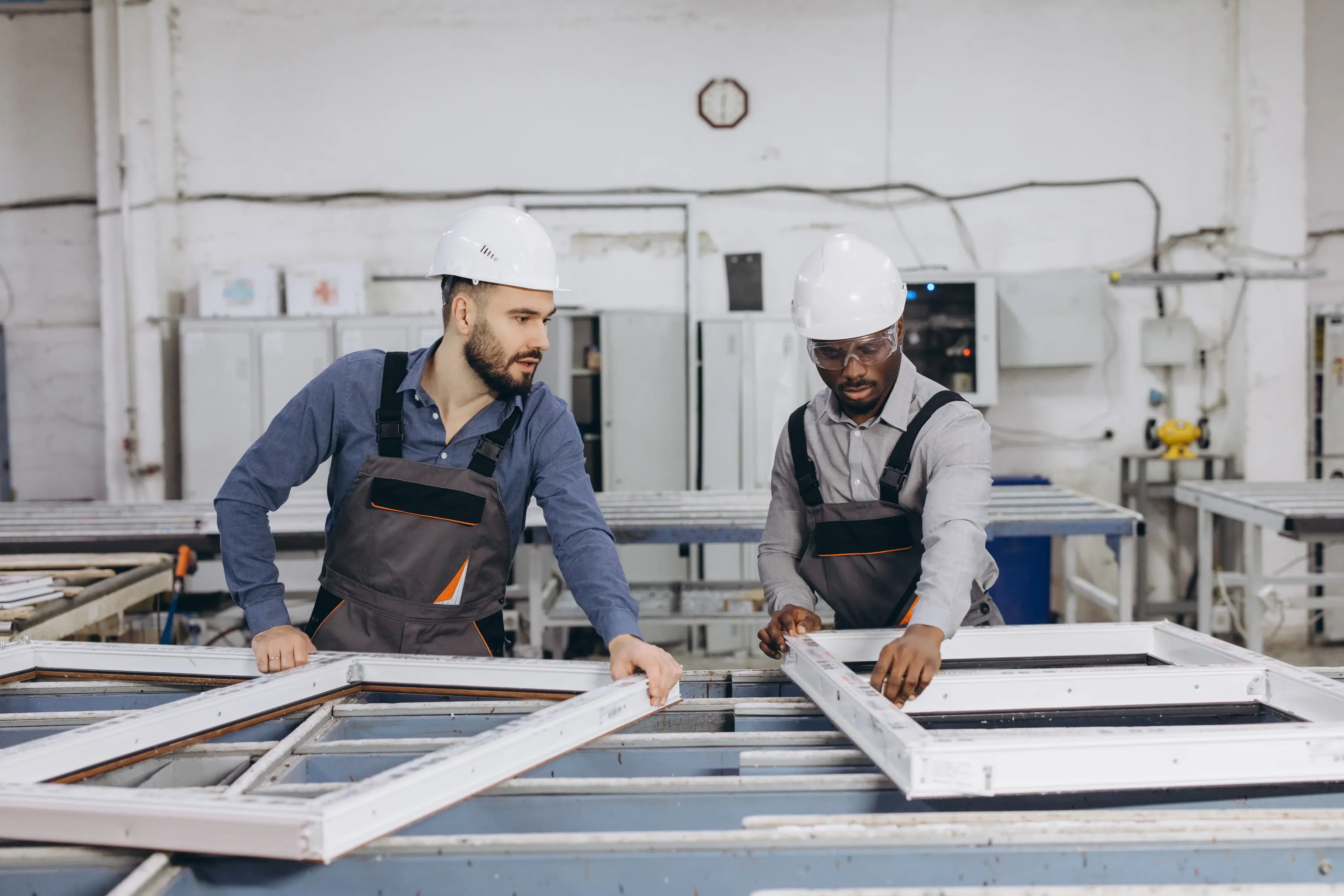 Two workers in hard hats inspecting window frames on a factory floor