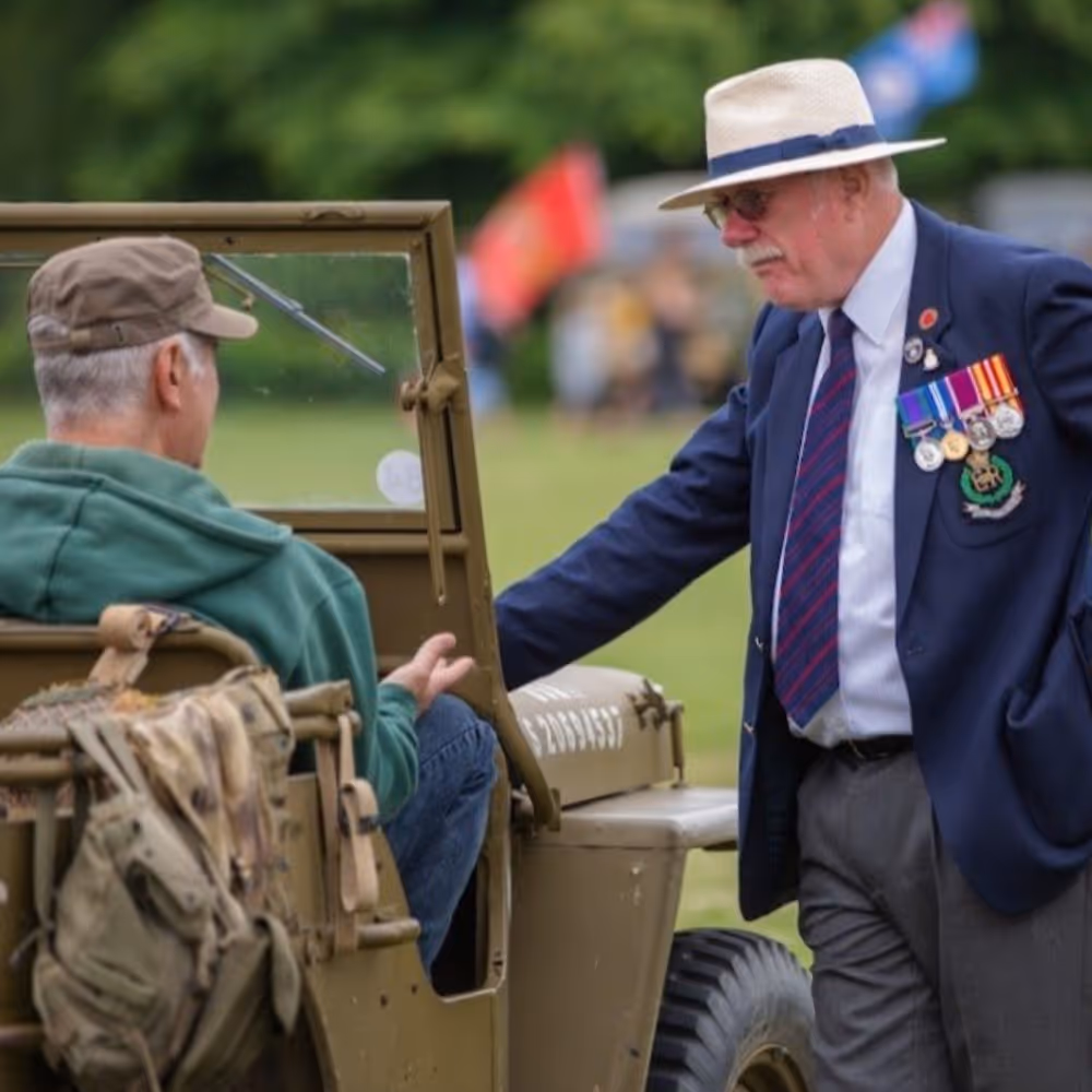 A man in a blue blazer adorned with war medals chatting to another man in an army jeep.