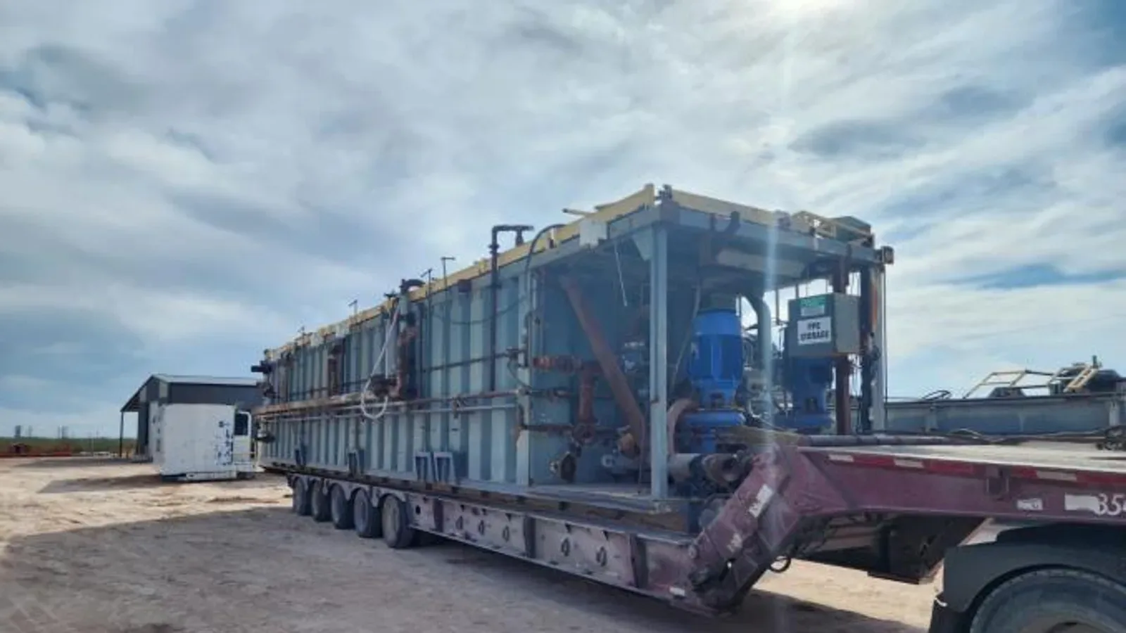 Large pneumatic dry bulk trailer with metal framework and piping parked in an industrial yard with cloudy sky overhead and industrial buildings visible