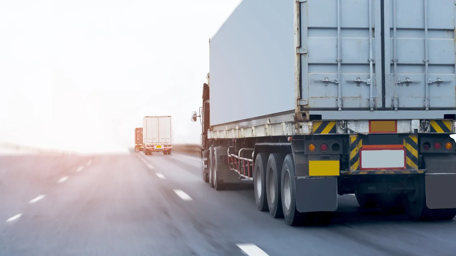 Several large cargo trucks traveling on a multi-lane highway in heavy fog conditions with reduced visibility and white lane markings visible.