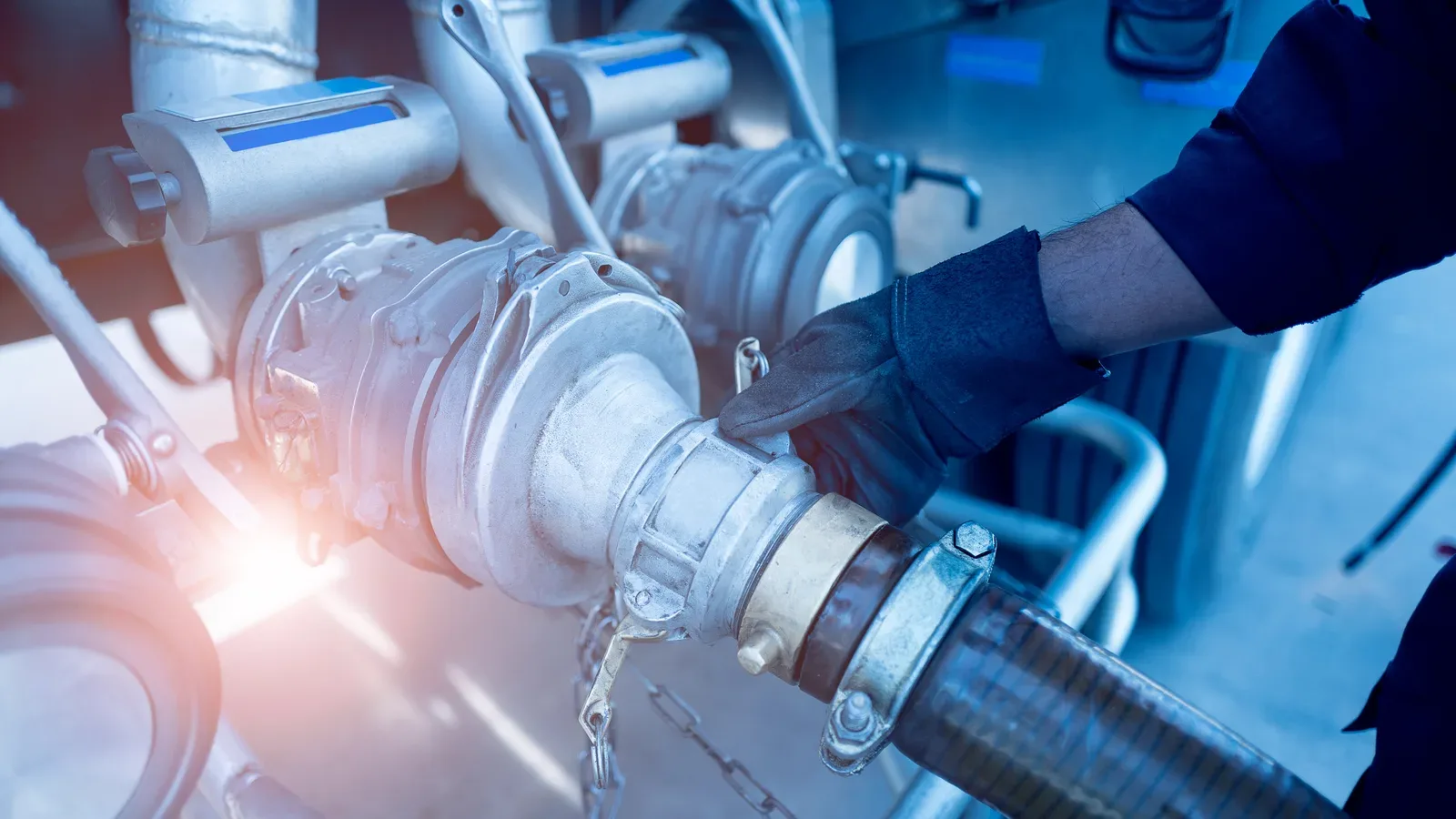 Technician in work gloves examines large industrial hose connections and valve fittings on tanker equipment. Blue and warm lighting effects visible.