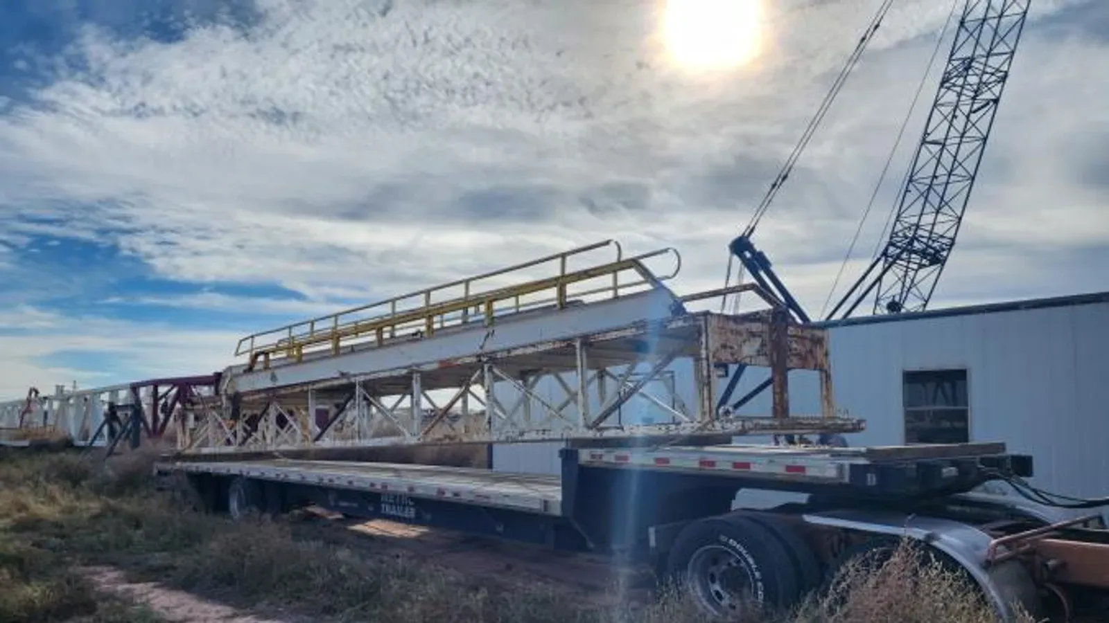 Flatbed trailer beneath a lattice tower crane in a heavy haul yard used for rig move equipment loading
