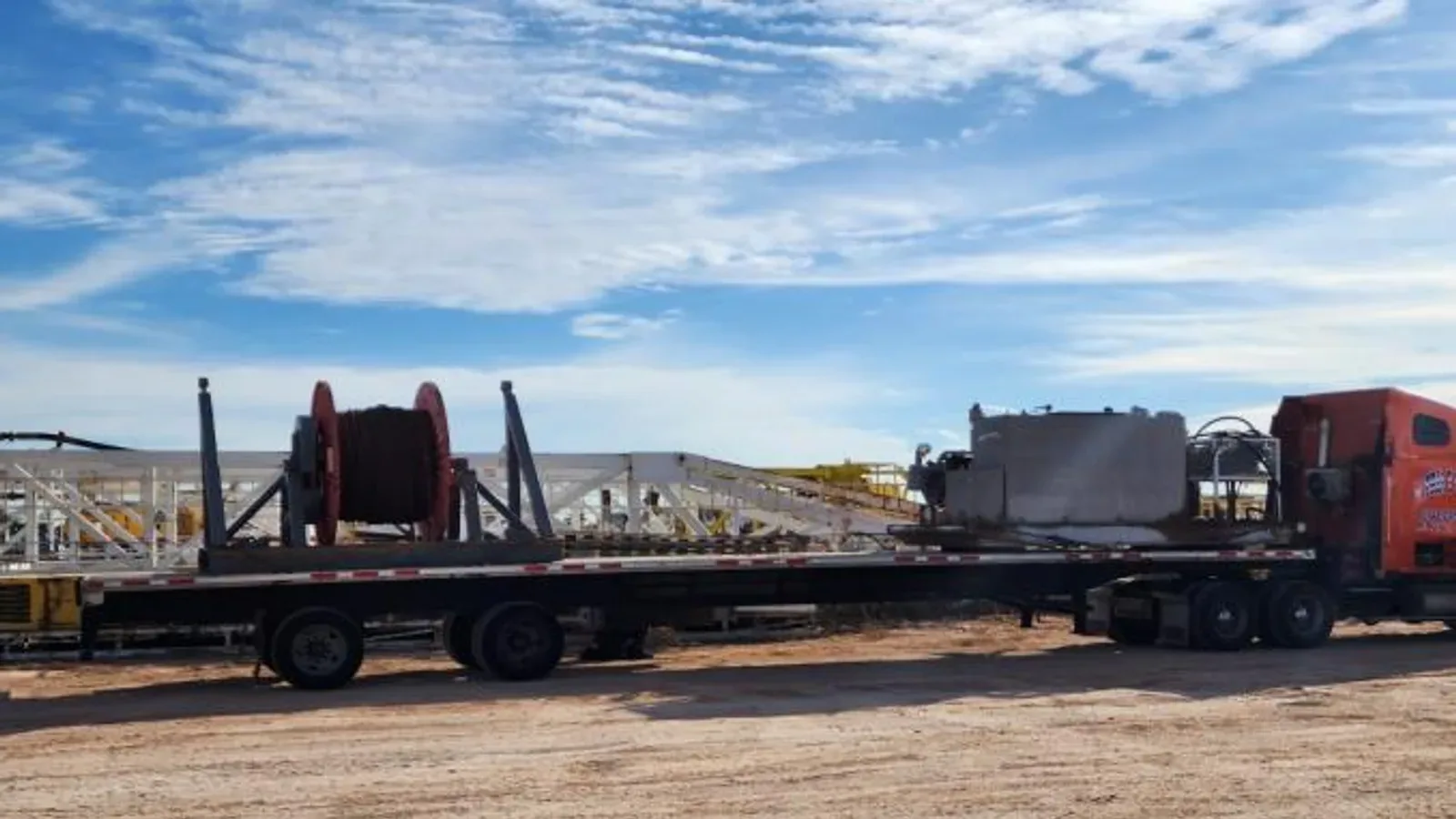 Red tractor unit pulling long flatbed trailer loaded with large spooled red cable at an industrial yard under clear blue sky