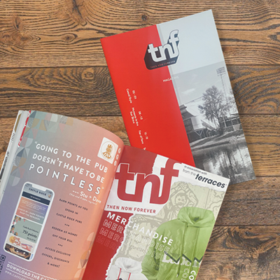Three colorful TNF-themed magazines fanned out on a wooden surface, featuring text and photos.