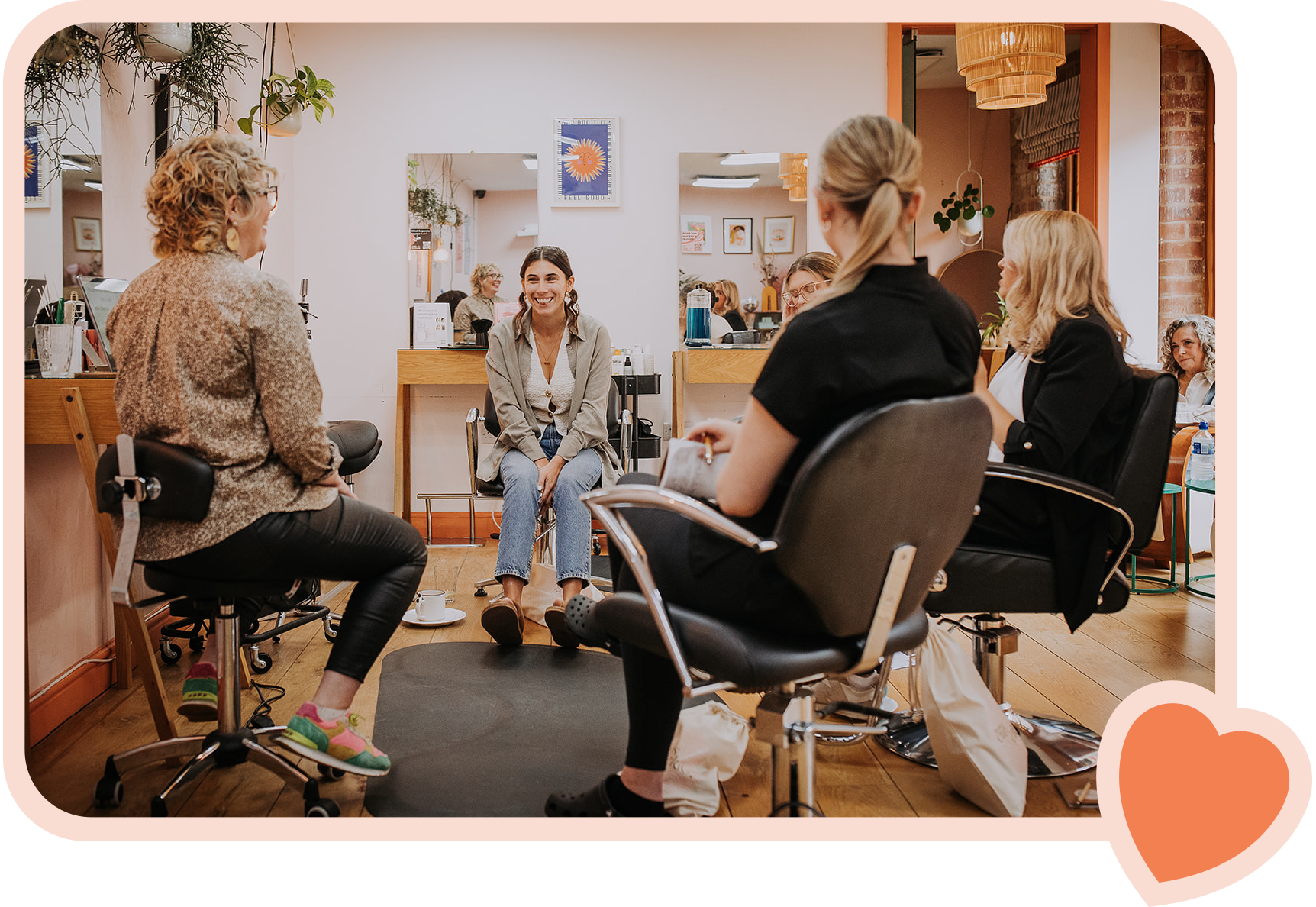 Five women seated in a salon studio, engaging in conversation and smiling, with mirrors behind them.