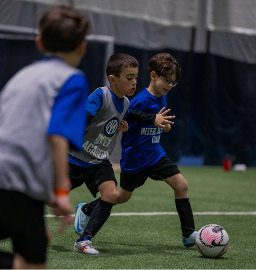 Two young boys in blue and gray sports jerseys playing indoor soccer, one controlling a white and pink soccer ball on green turf.