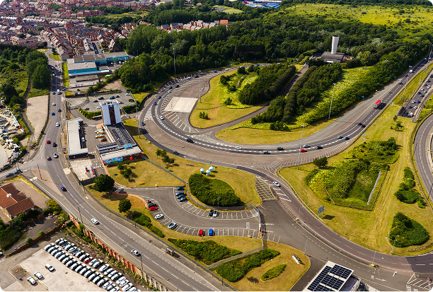 Aerial view of a highway cloverleaf interchange surrounded by green grassy areas, trees, parked cars, and nearby buildings leading into a residential neighborhood.