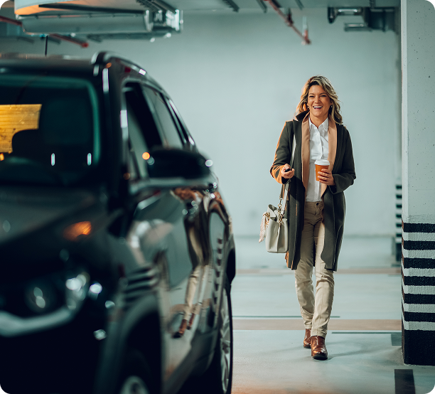 Smiling woman walking in a parking garage carrying a coffee cup and keys, approaching a black SUV.