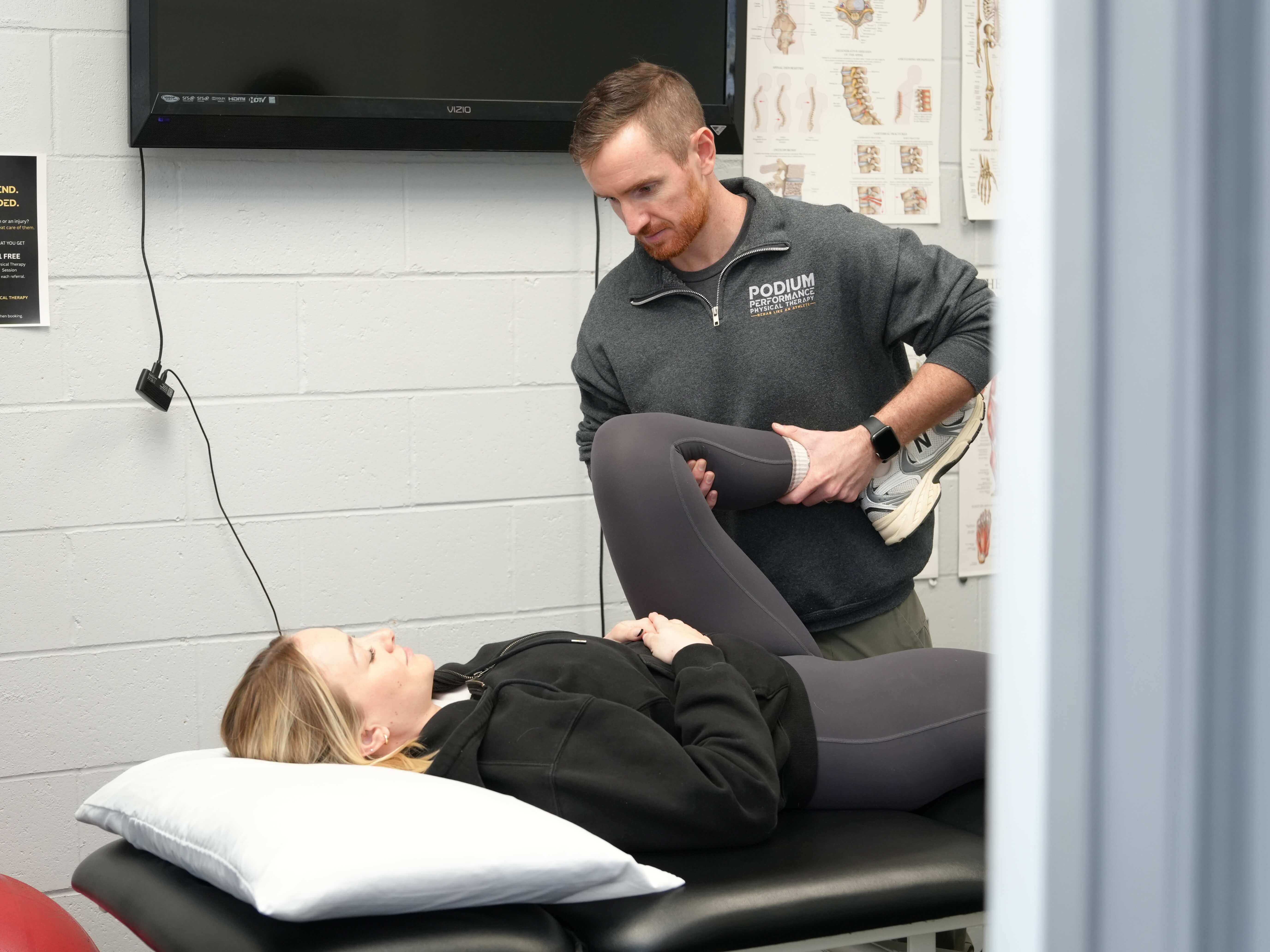 Physical therapist holding and stretching a female patient's bent leg on a treatment table in a therapy room.