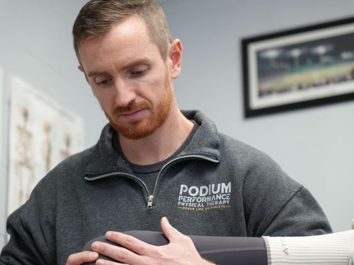 Therapist in Podium Performance Physical Therapy jacket examining a patient's leg in a clinical setting.