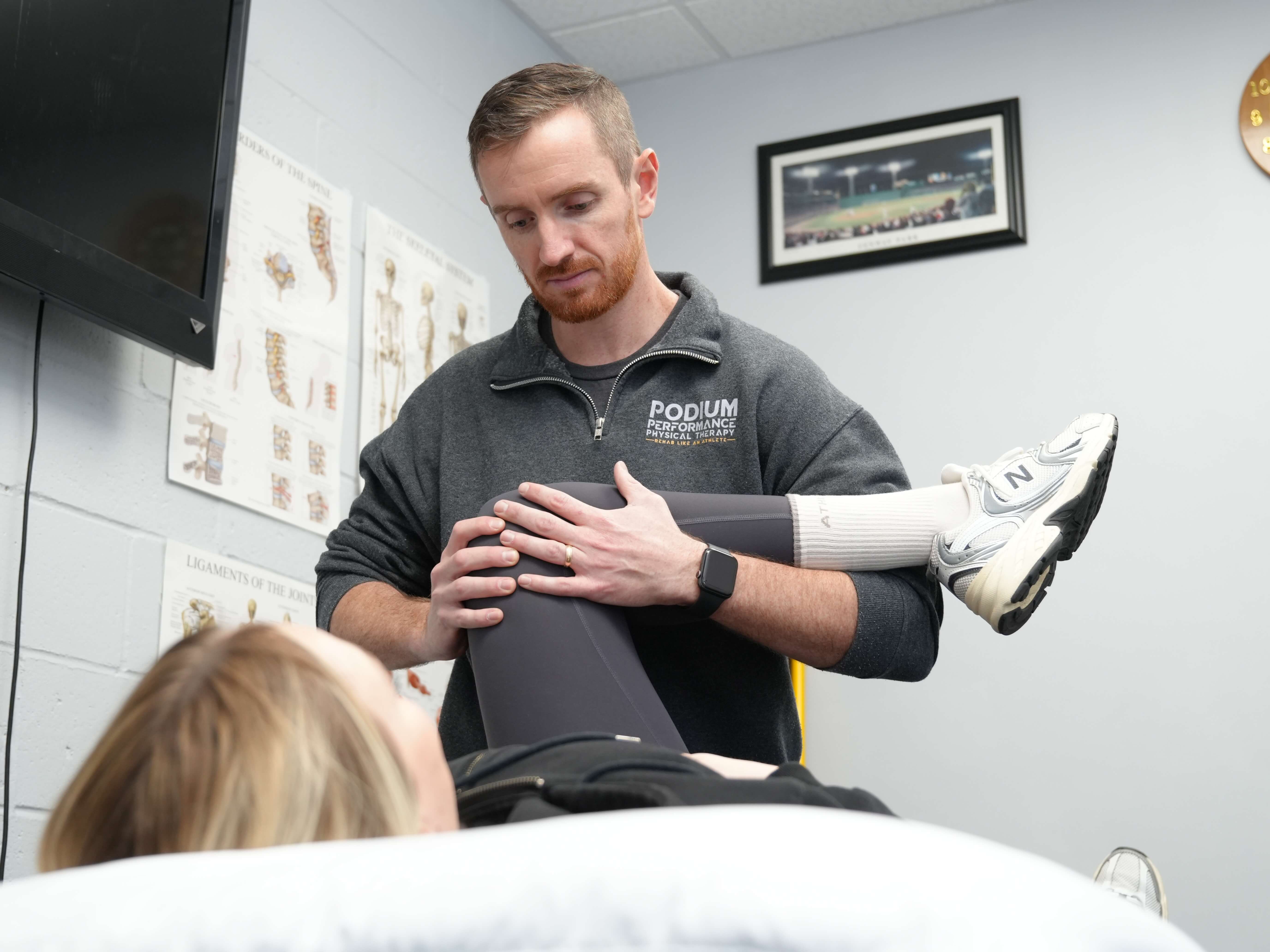 Physical therapist in Podium Performance sweatshirt assisting a patient by stretching her bent knee in a clinic room.
