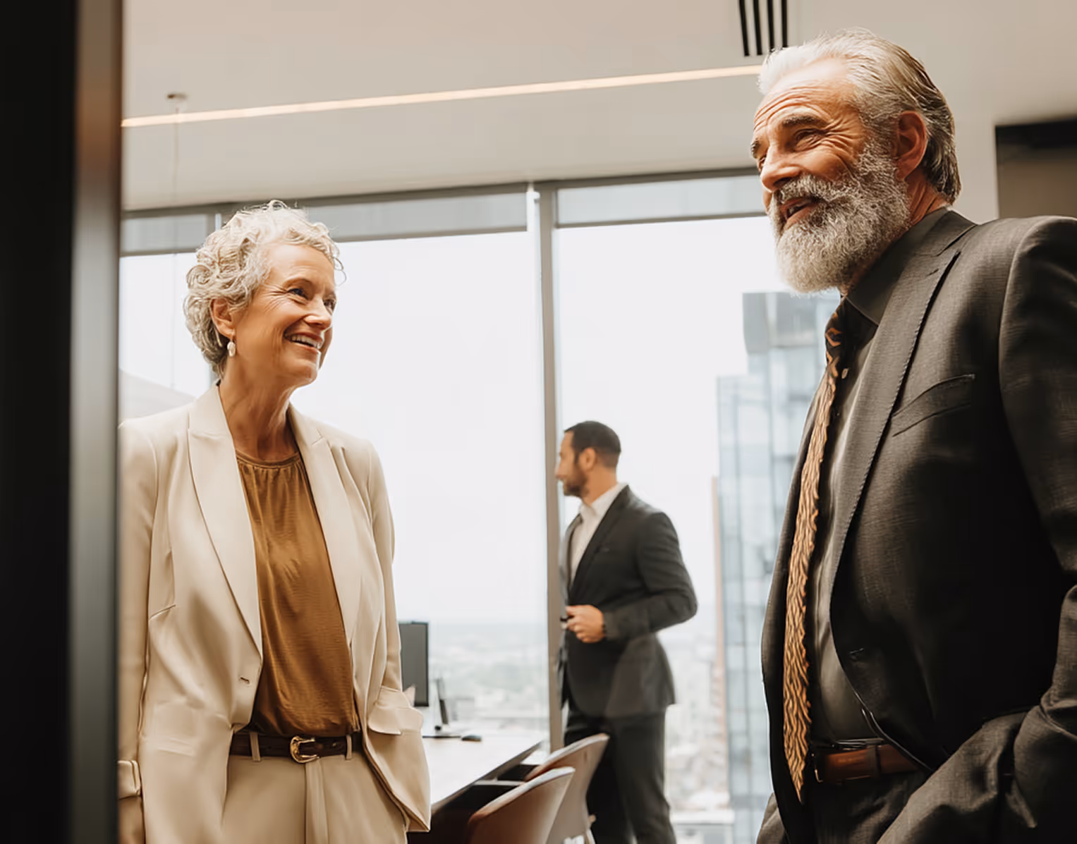 Two older professionals, a woman and a bearded man, smiling and talking in a bright office with a younger man standing in the background near a window.