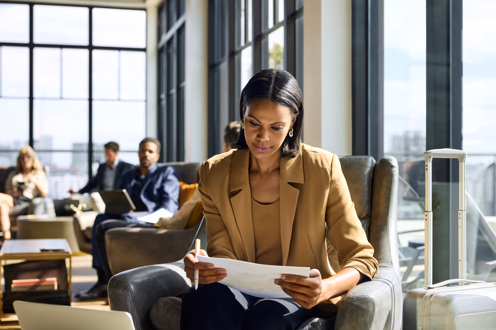 Businesswoman in a tan blazer reviewing documents while seated in a modern lounge with large windows and a suitcase nearby.