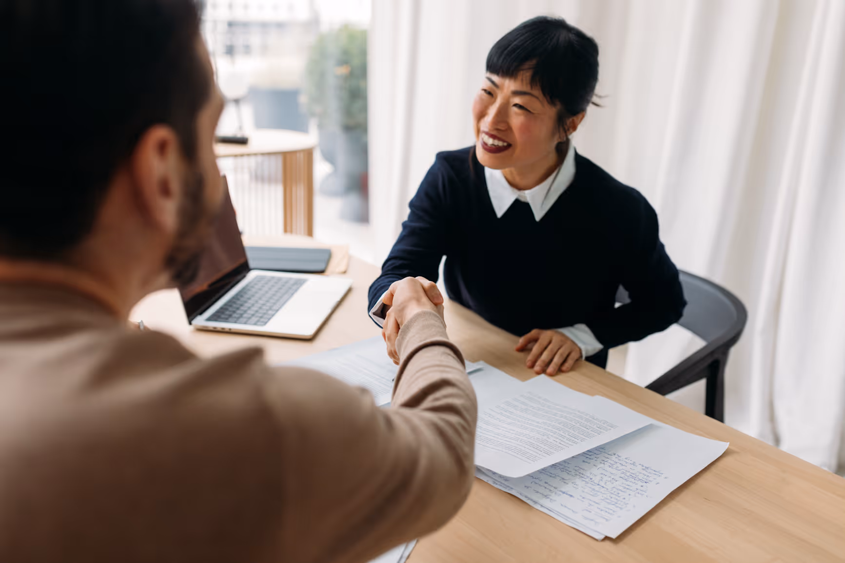 Two people shaking hands over a desk with documents and a laptop, smiling during a meeting.
