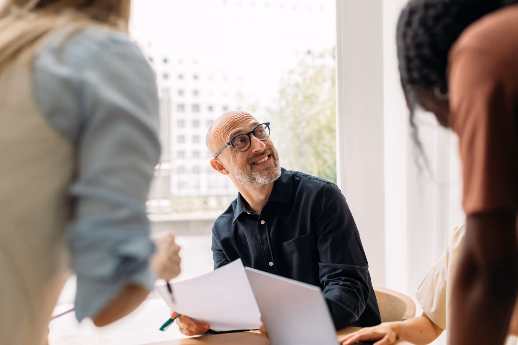 Smiling bald man with glasses holding papers, looking up during a meeting with colleagues.