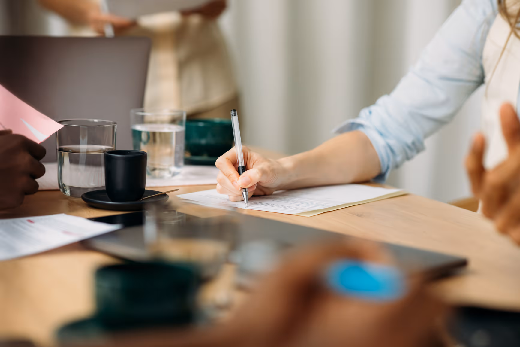 Person signing a document on a desk with a laptop, glasses of water, and coffee cups nearby.