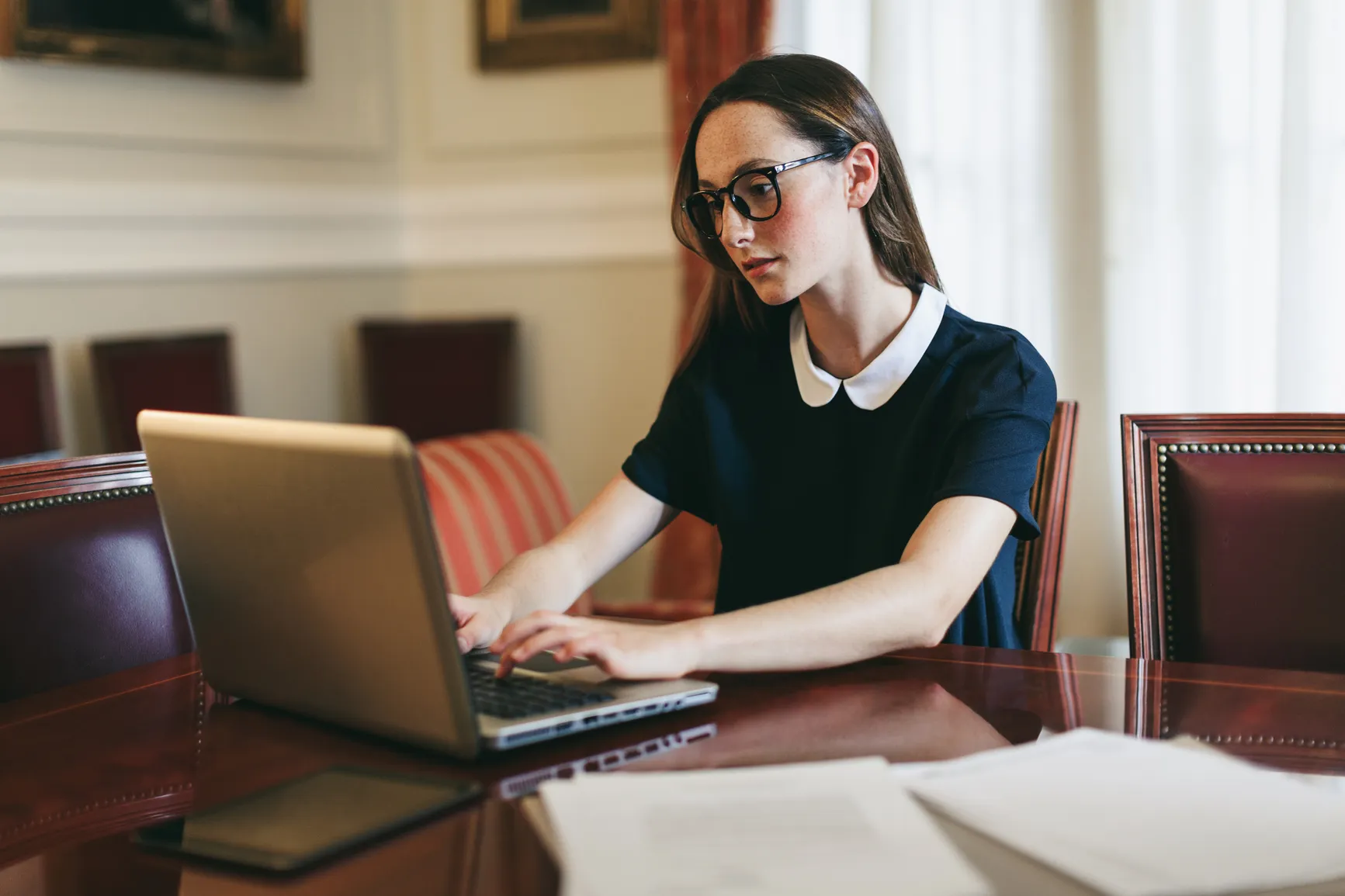 Young woman wearing glasses typing on a laptop at a polished wooden table in a formal room.